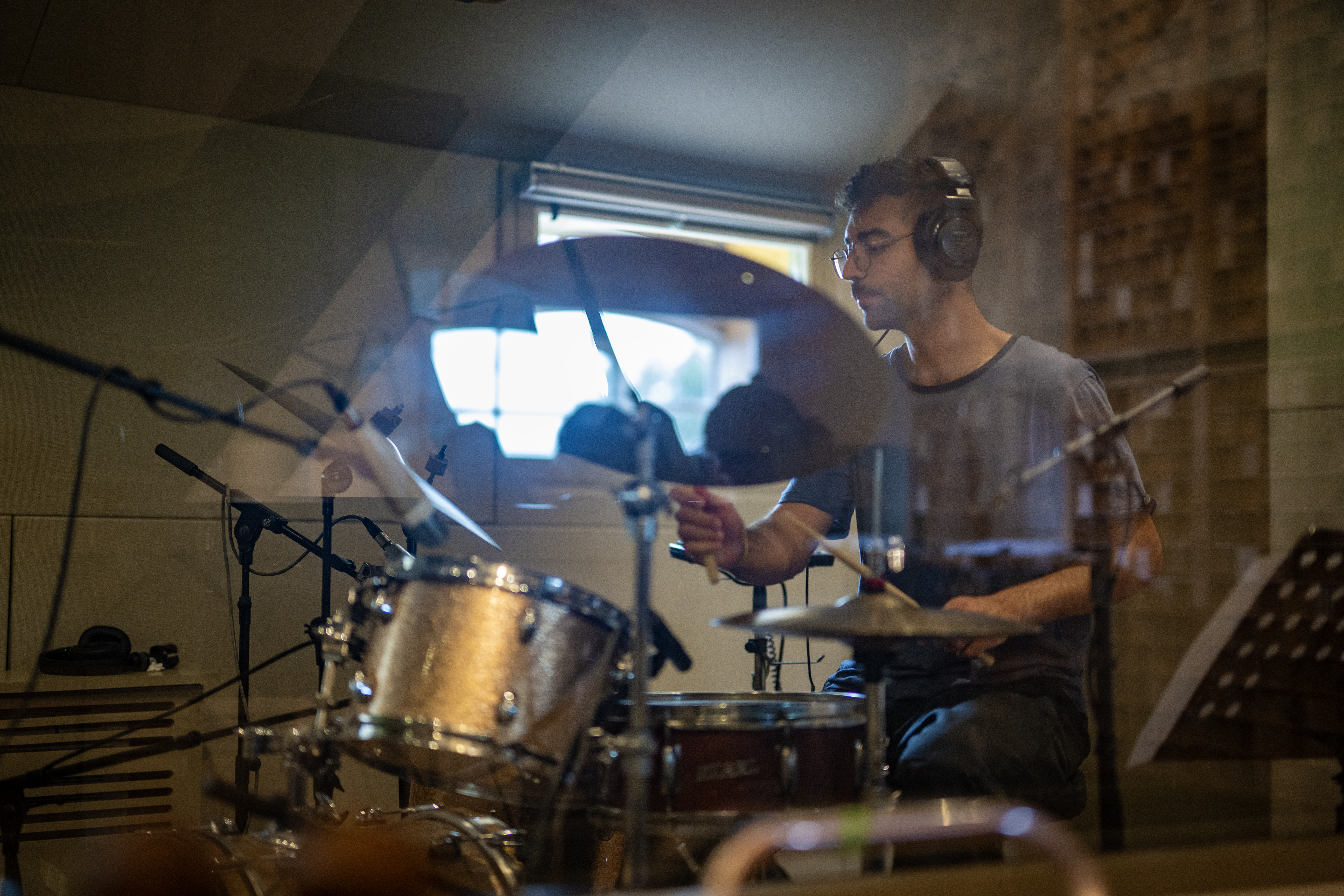 The drummer playing during a recording session, viewed through the studio glass.