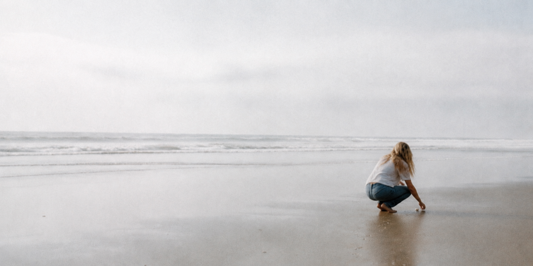 A woman at the shoreline reaching toward something in the sand.