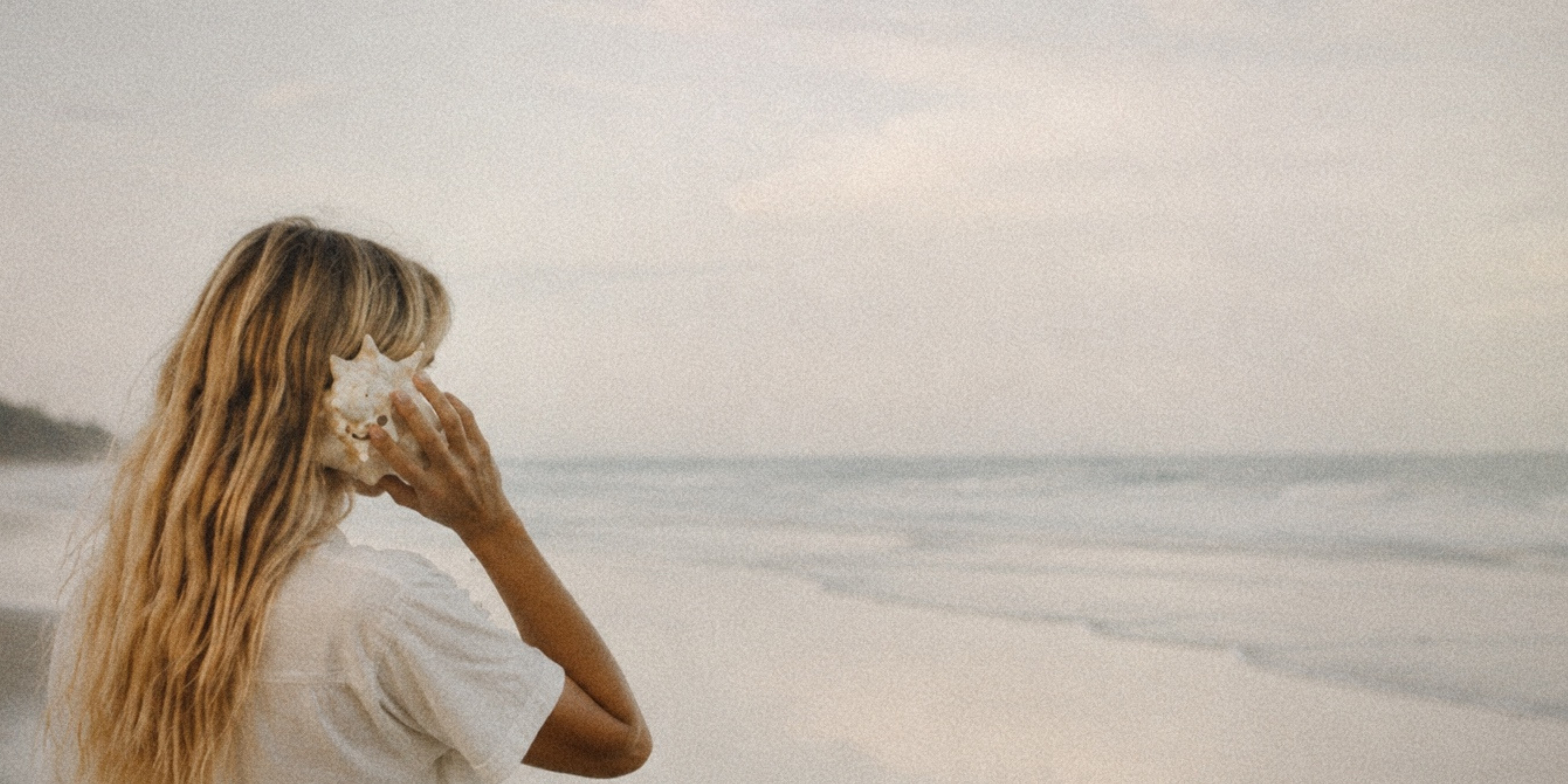A woman holding a seashell to her ear, listening, beside the ocean