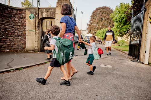 A parent walking two children with backpacks towards the school gate