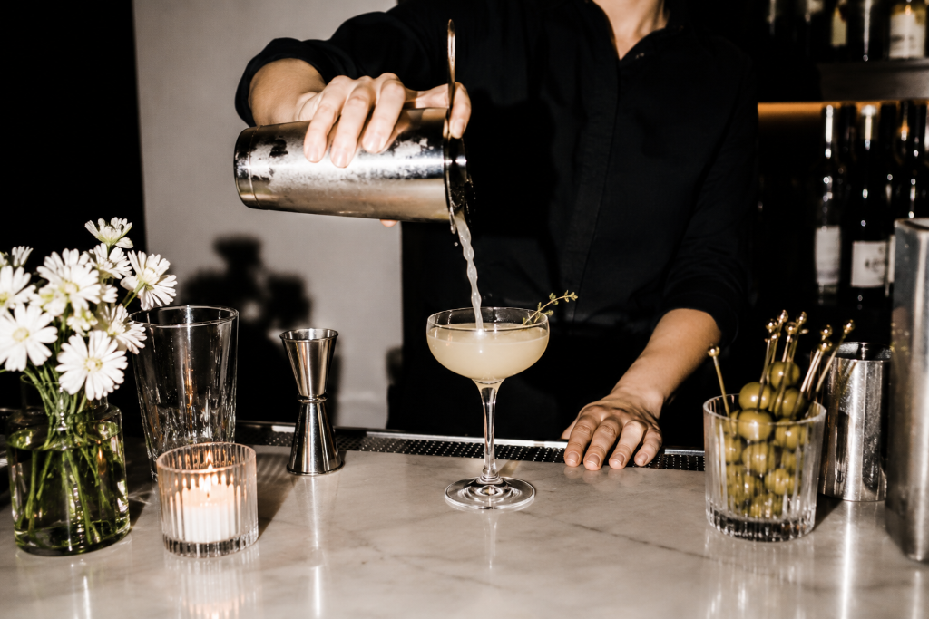 A bartender pouring a cocktail into a coupe glass on a marble bar
