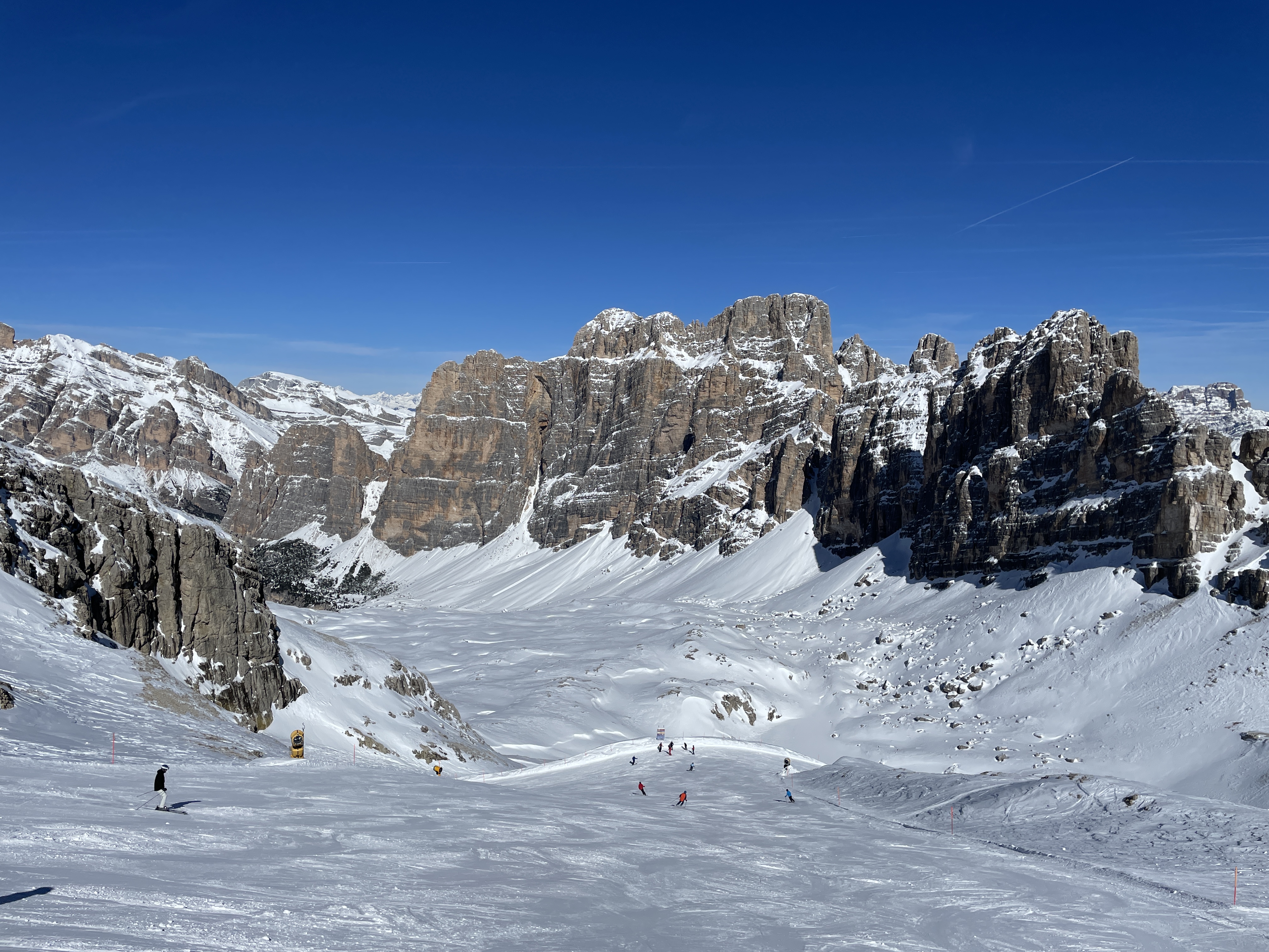 Skiers on a wide piste below jagged rock towers