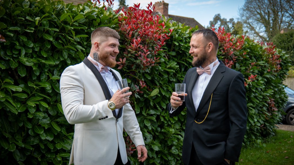 Two men holding drinks and laughing in front of a hedge