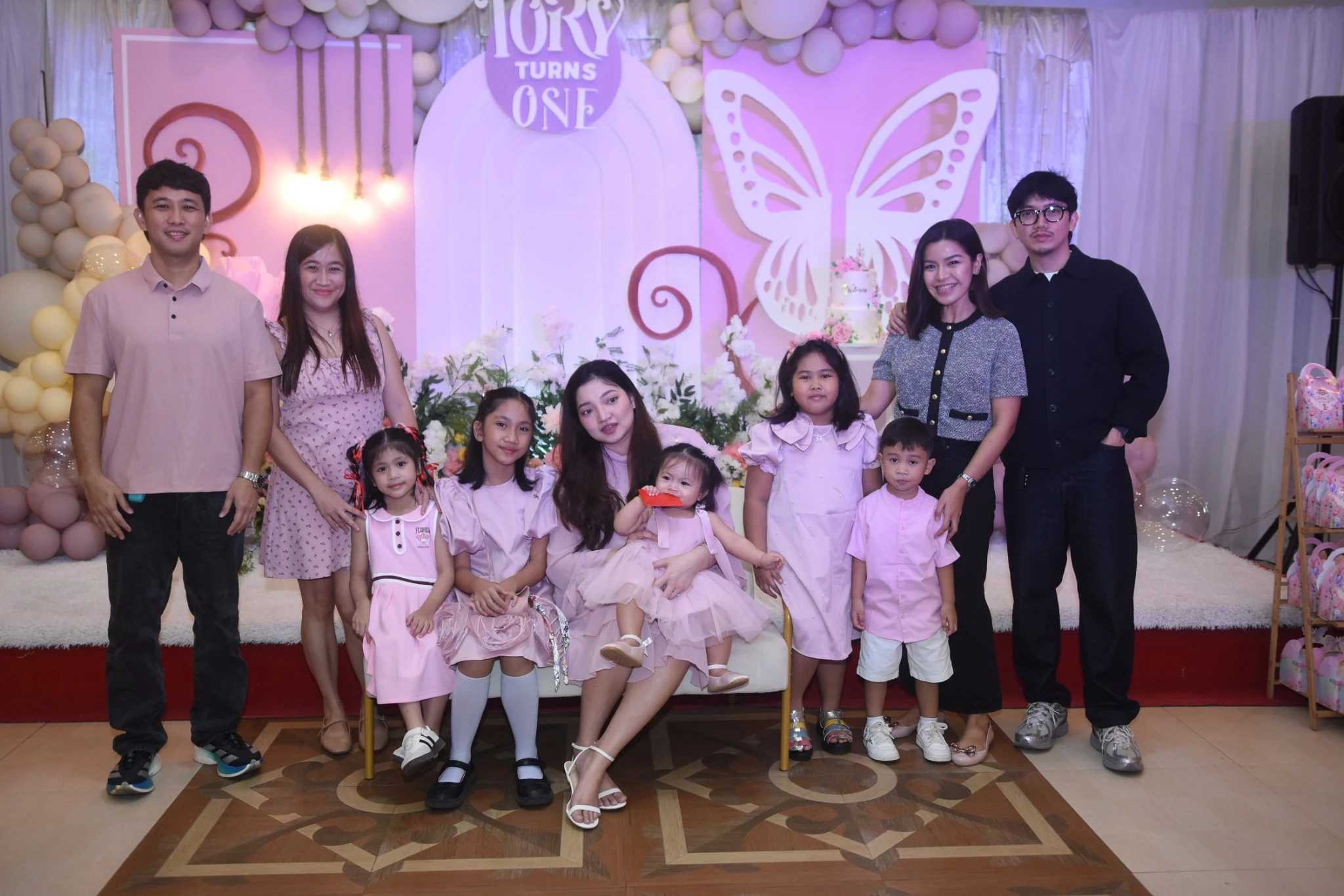 Children and adults gather around the birthday child for a group portrait beside the butterfly decorations.