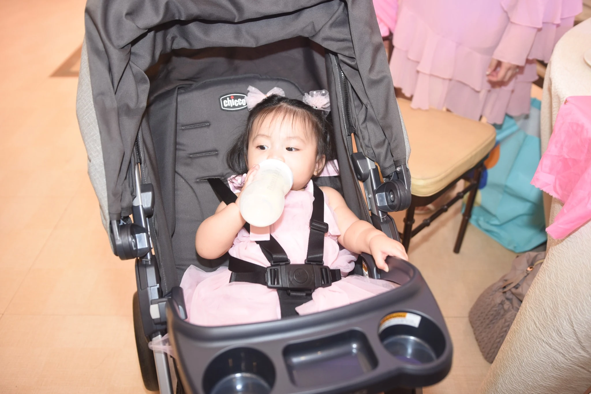 A child in a pink dress drinks from a bottle while sitting in a stroller at the party.