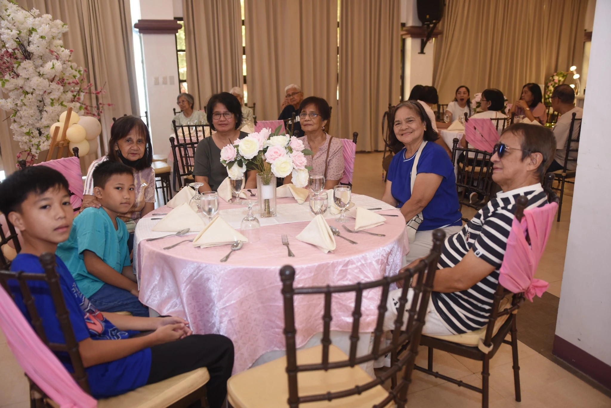Guests and children sit at a round table with pink linens and a floral centerpiece.