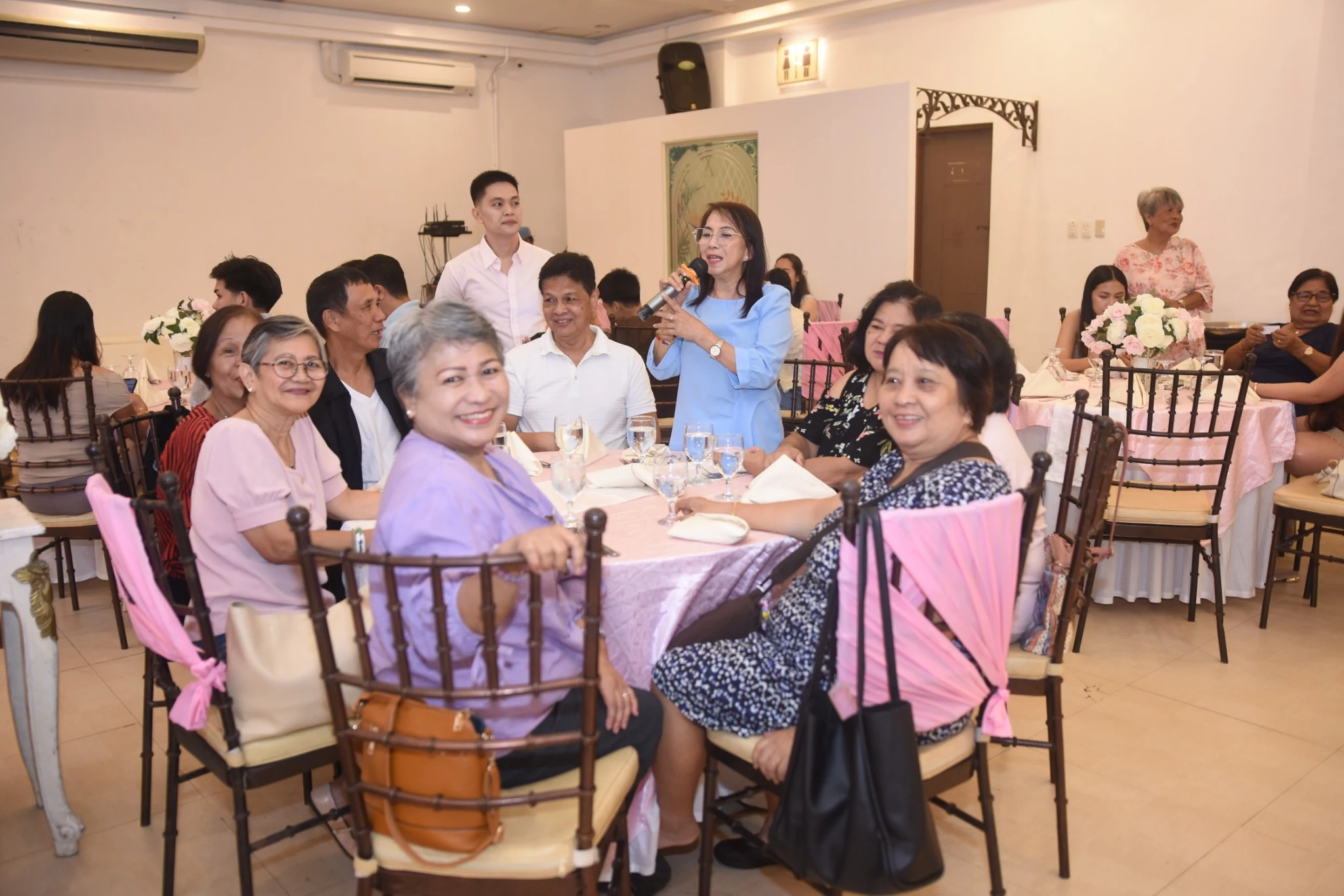 A standing guest holds a microphone while other guests smile around the birthday reception tables.