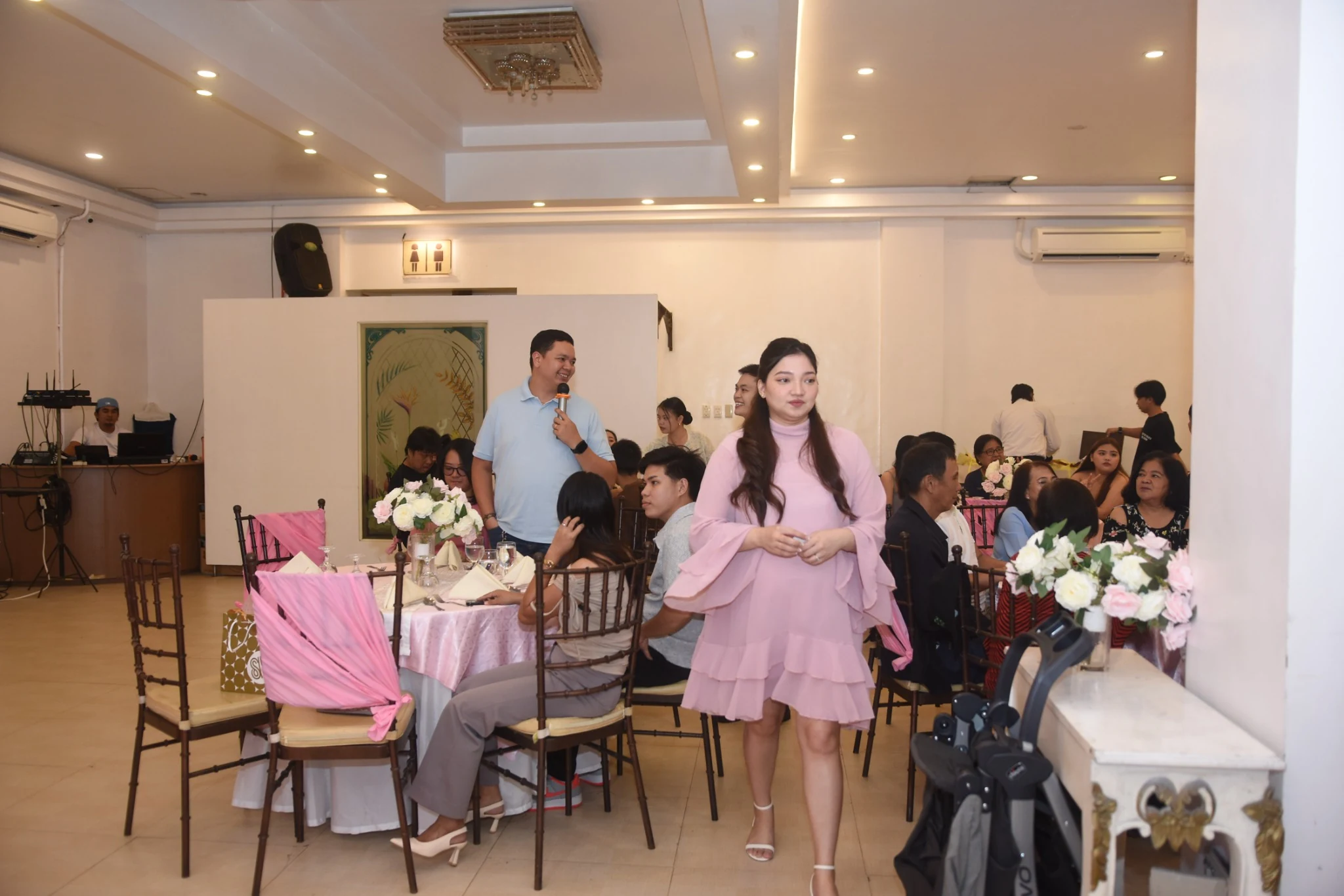 A guest in a pink dress walks between reception tables while another guest holds a microphone.