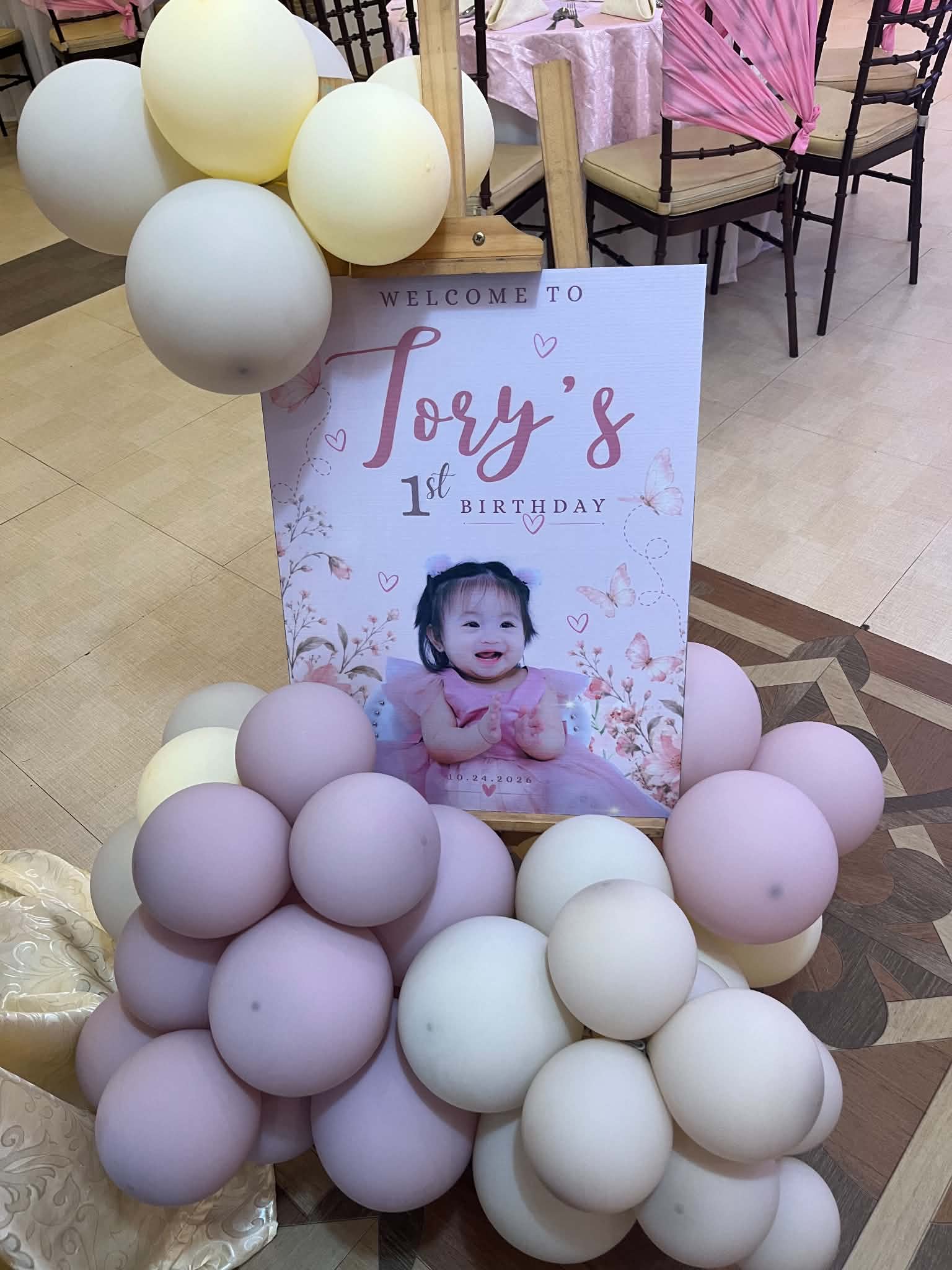 Tory's first-birthday welcome sign with her smiling photograph, surrounded by cream and lilac balloons.