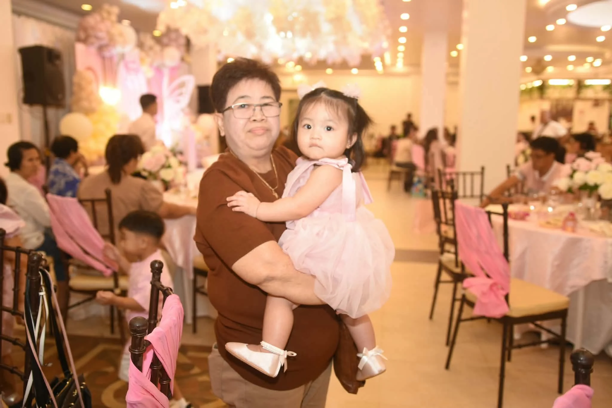 A guest holds a child in a pink dress in the warmly lit birthday reception room.