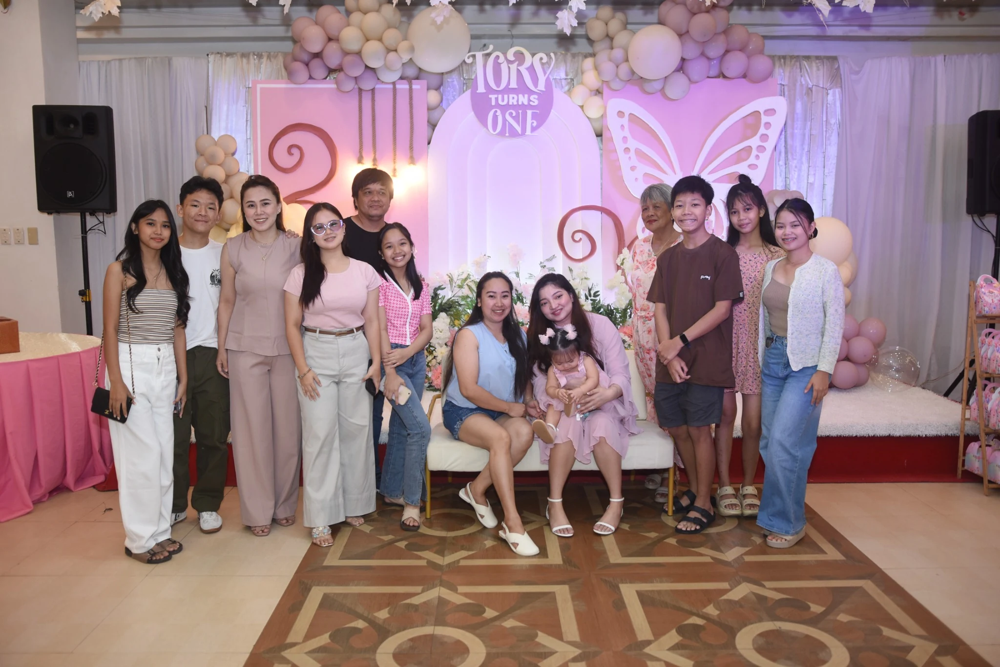 A large group of adults and children gathers for a birthday portrait in front of the pastel stage.