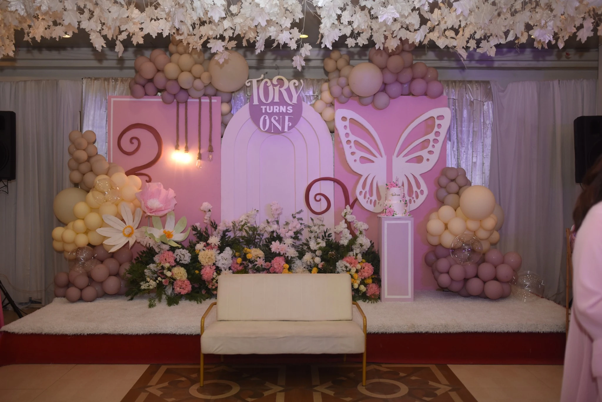 A pastel pink and lilac birthday backdrop with balloons, flowers, a butterfly decoration, and a Tory Turns One sign.