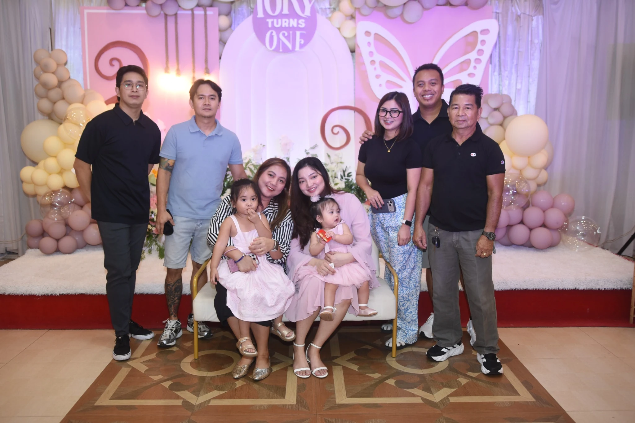 Adults and children pose together in front of the pastel first-birthday backdrop.