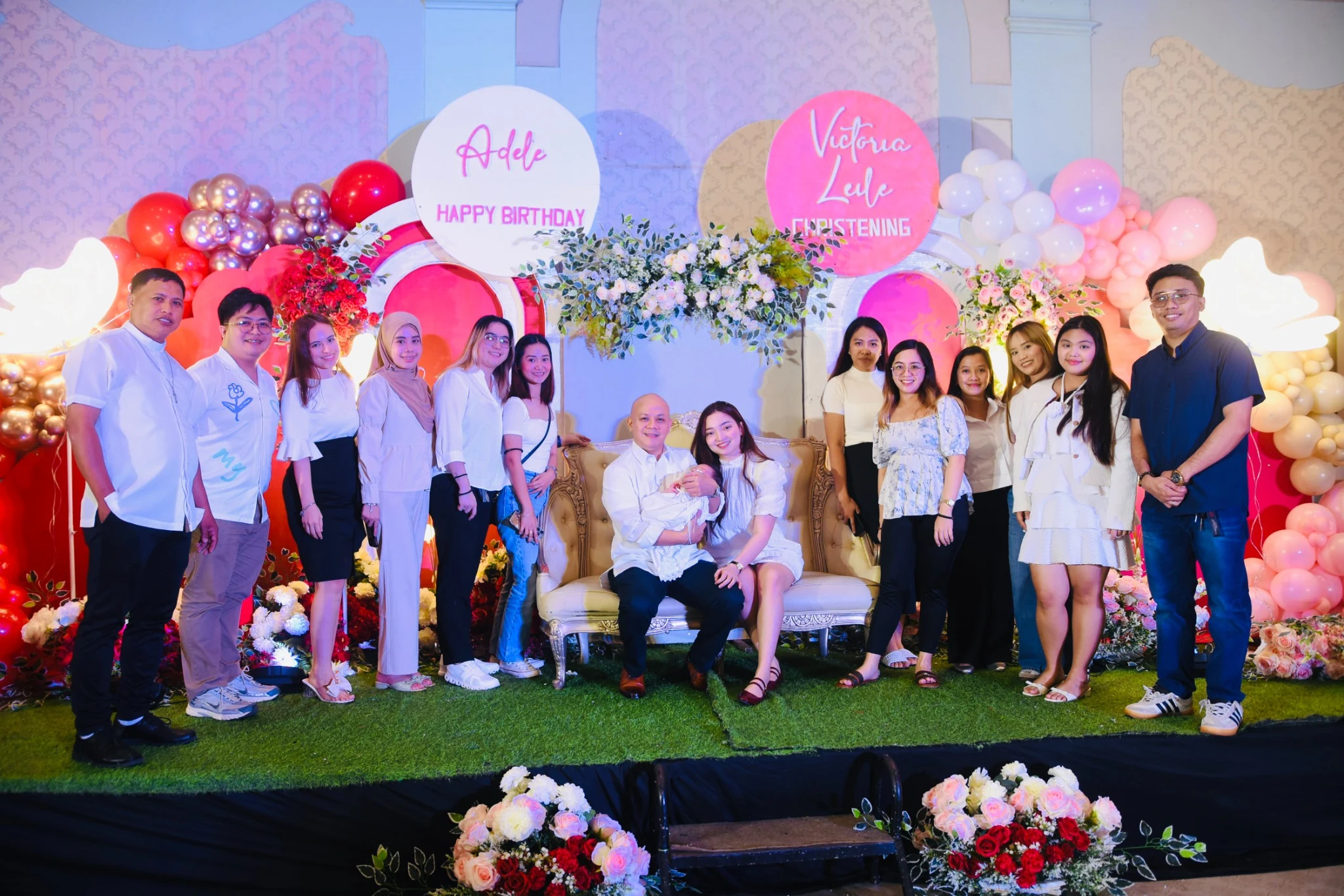 A large group poses around the seated adults and baby on the decorated stage.
