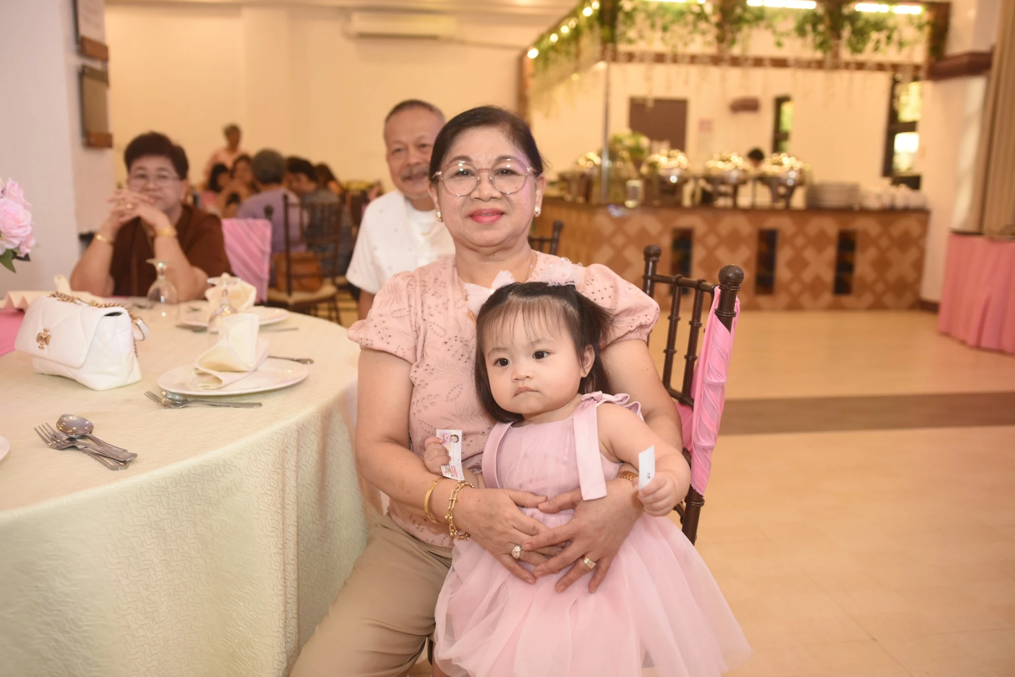 A child in a pink birthday dress sits on a guest's lap beside the celebration table.