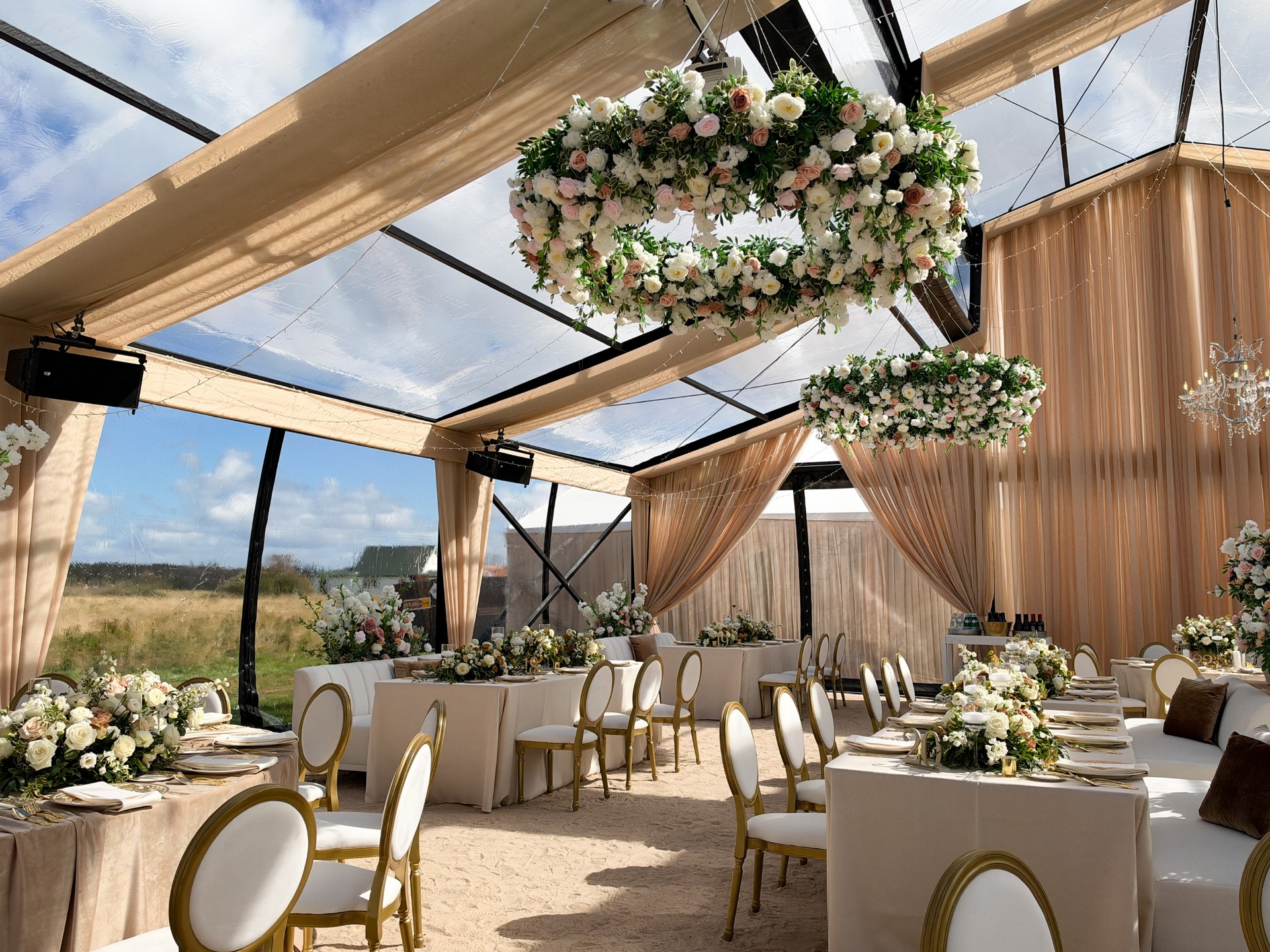 Wedding reception with linen-covered tables, gold chairs and hanging floral arrangements beneath a draped glass ceiling