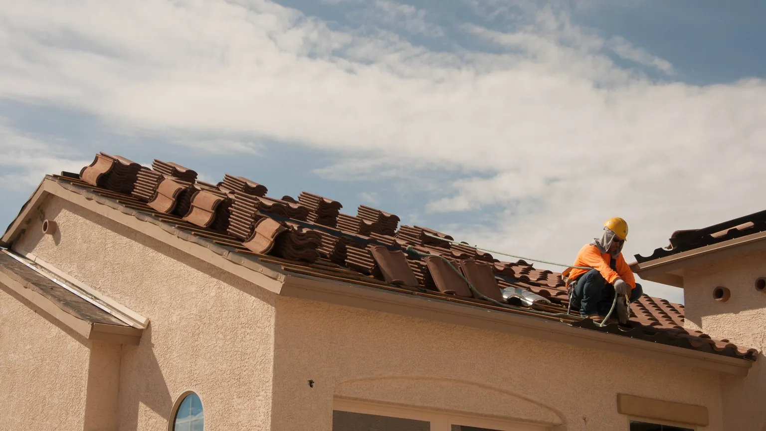 A roofer wearing a hard hat works beside stacks of curved roof tiles on a sloped residential roof.