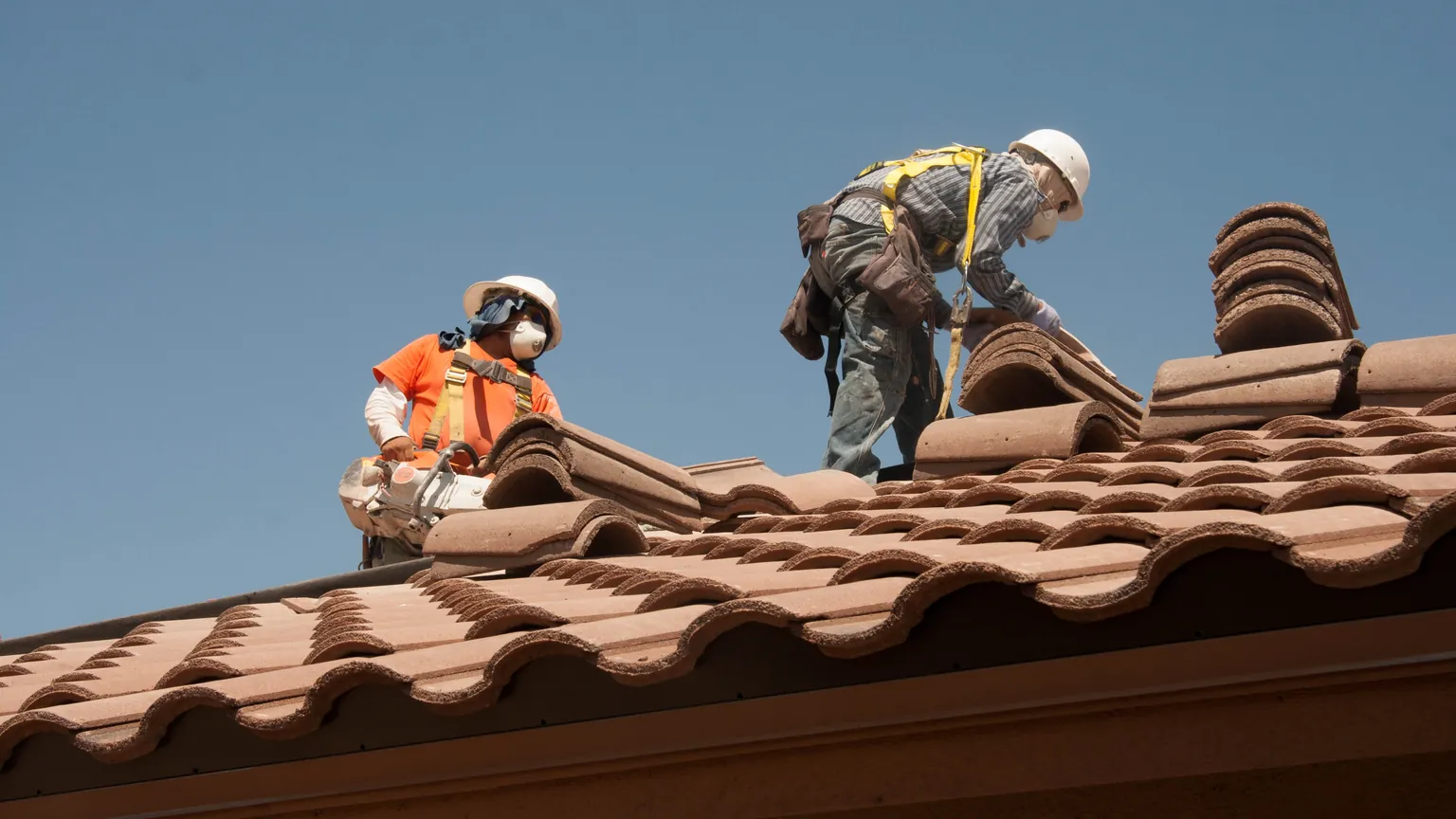 Two roofers wearing hard hats, respirators and safety harnesses handle curved tiles on a sloped roof.