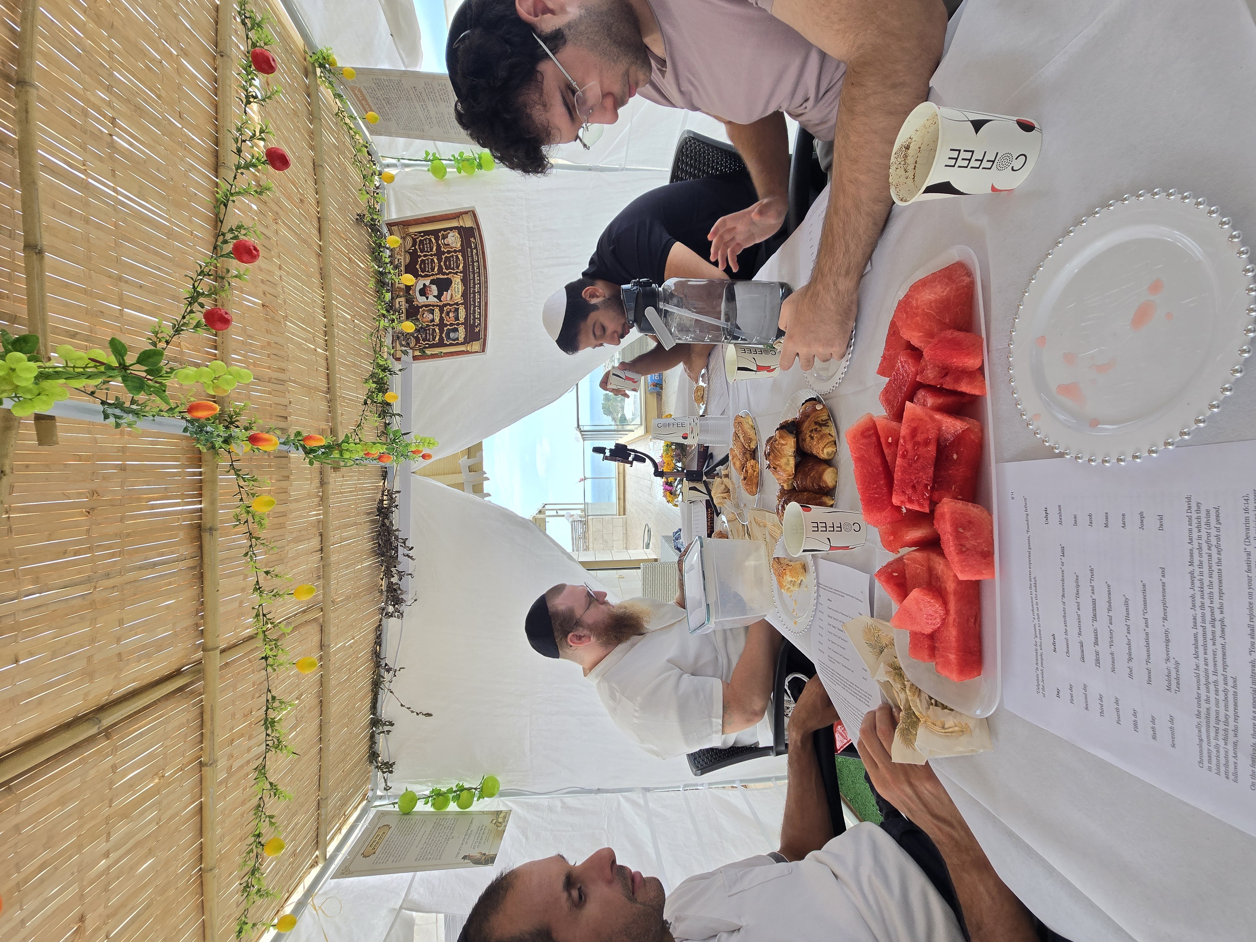 Four chairs filled, a watermelon cut open, and the week's page spread on the table — nobody is in a hurry. A Shabbat that starts as a tablecloth and ends as family.
