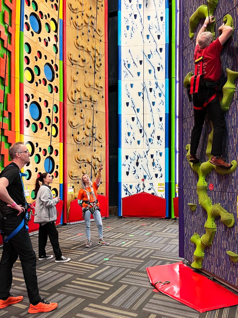 Corporate group participants at the Clip 'n Climb walls, with staff instructor guiding a man scaling the colorful climbing wall at Allegan Event