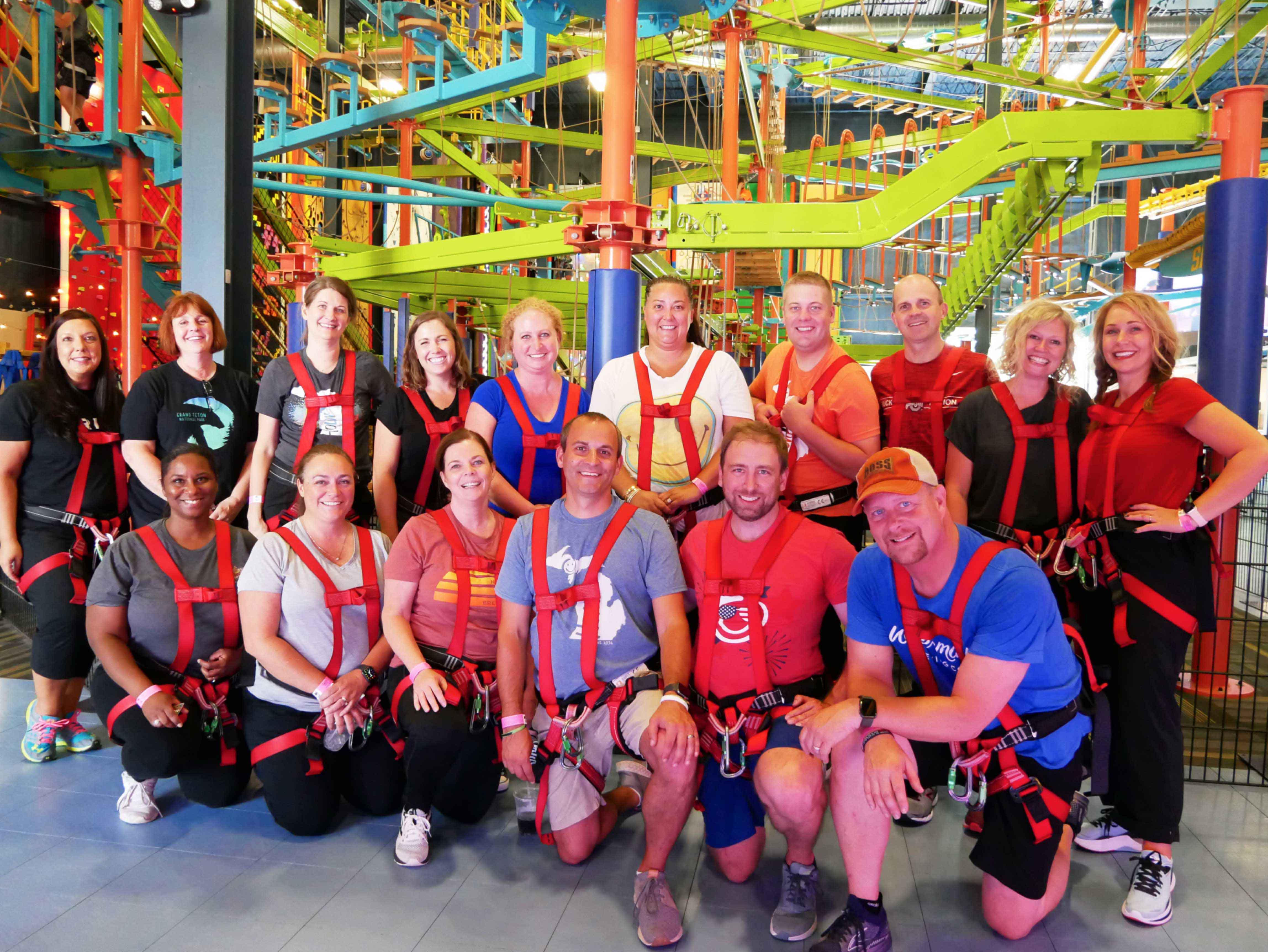 Corporate team group photo smiling and cheering in harnesses in front of the Sky Trail ropes course at Allegan Event
