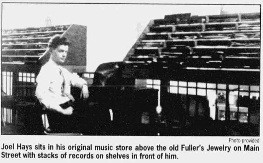 Black and white newspaper photo of Joel Hays seated in his music store surrounded by tiered record shelves