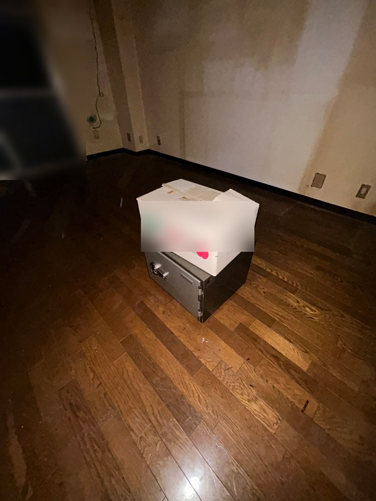 フローリングの洋室。ほかに物のない床の中央に、金庫が1つと、その上に段ボール箱が1つ置かれている