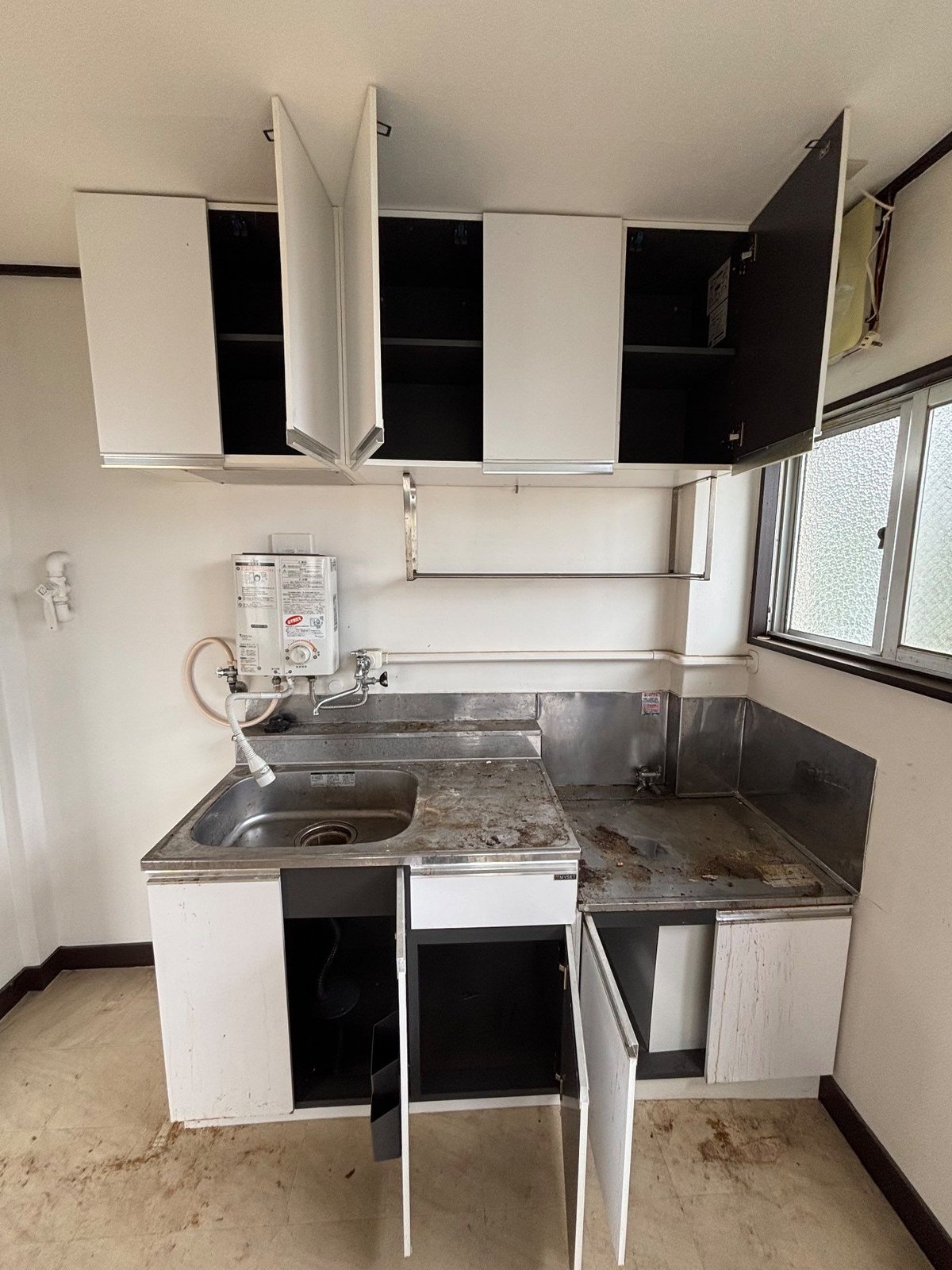 作業後の2階の台所。吊り戸棚と流し台の下の扉がすべて開いていて、中は空になっている。壁には小型の湯沸かし器が付いている