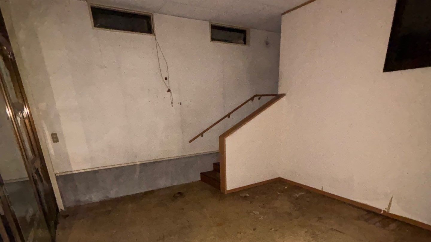 家財を運び出した後の部屋。右手に上へ続く階段と手すり、奥の壁に高い位置の窓が2つあり、床には物が残っていない