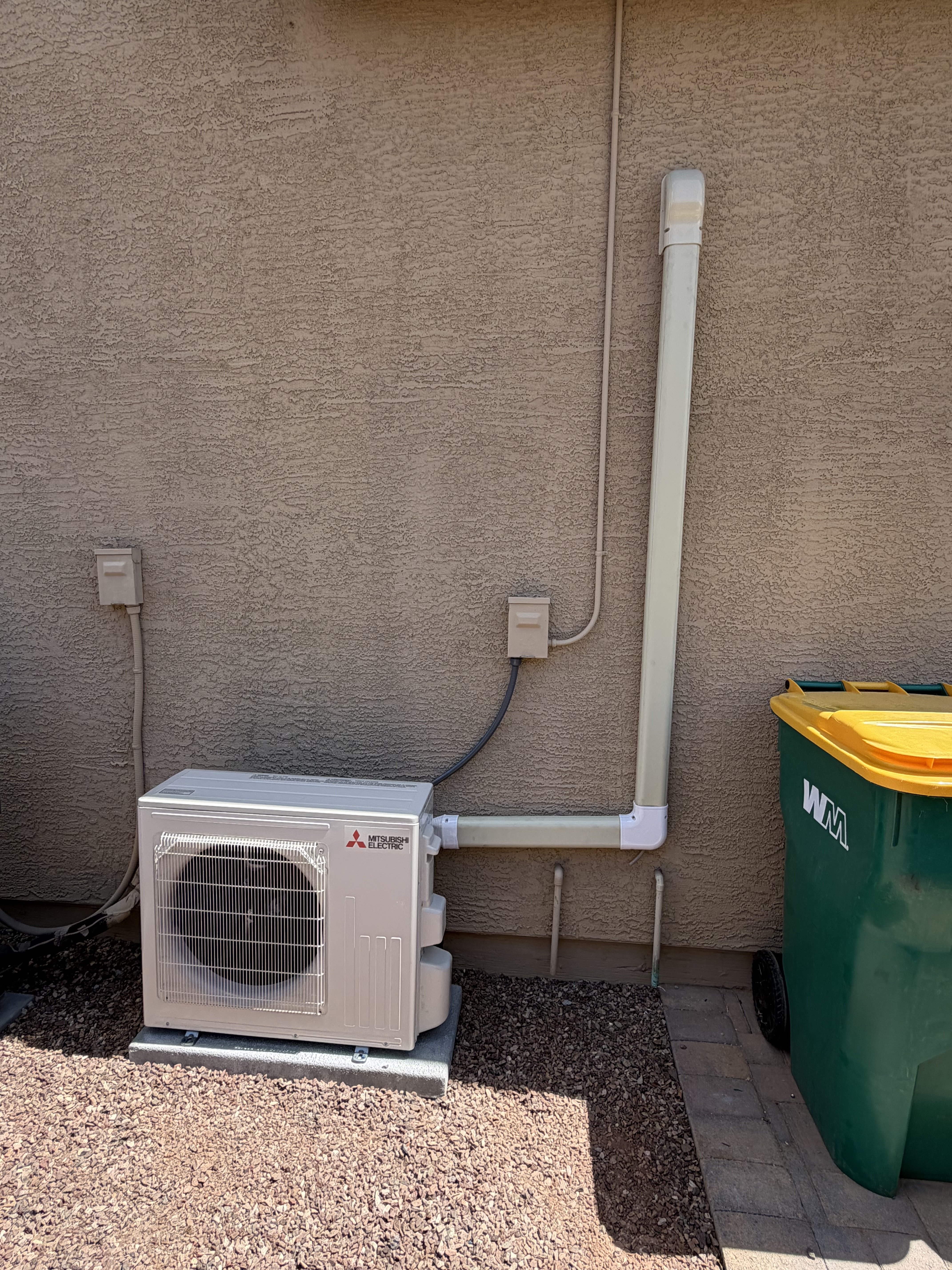 Outdoor Mitsubishi Electric mini-split condenser unit installed on a concrete pad against a stucco house wall, with a white protective line set cover and an electrical disconnect box.
