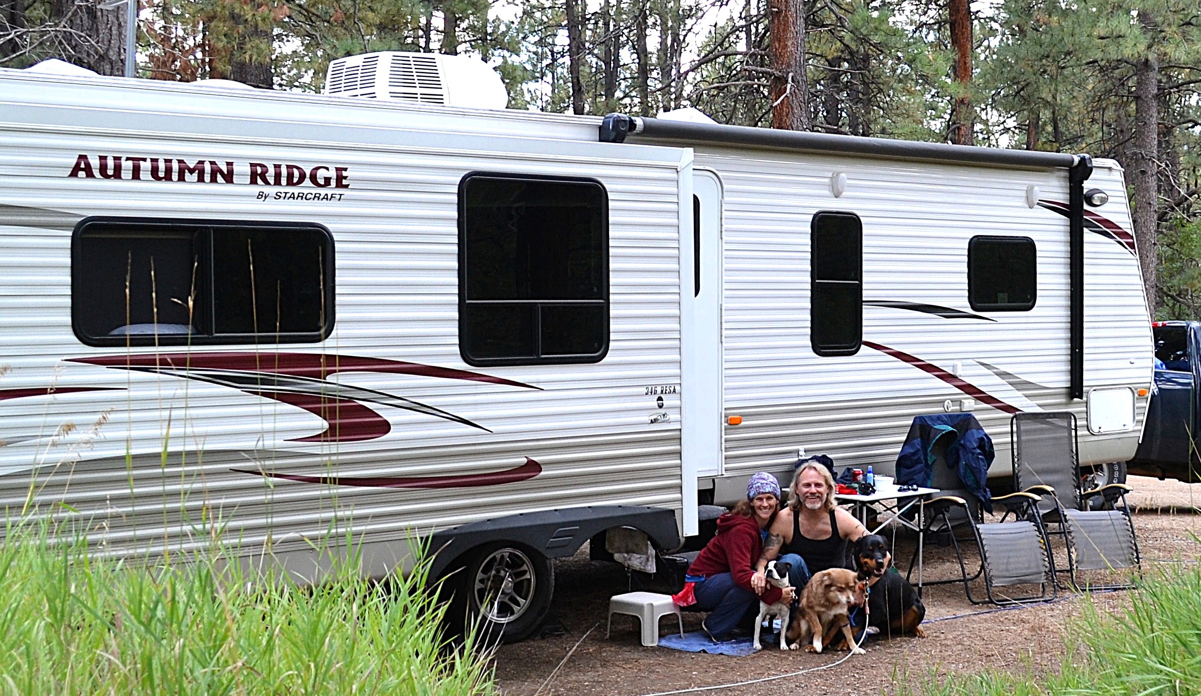 Tara and John sitting in camp chairs with their dogs next to their Starcraft Autumn Ridge travel trailer at a wooded campsite