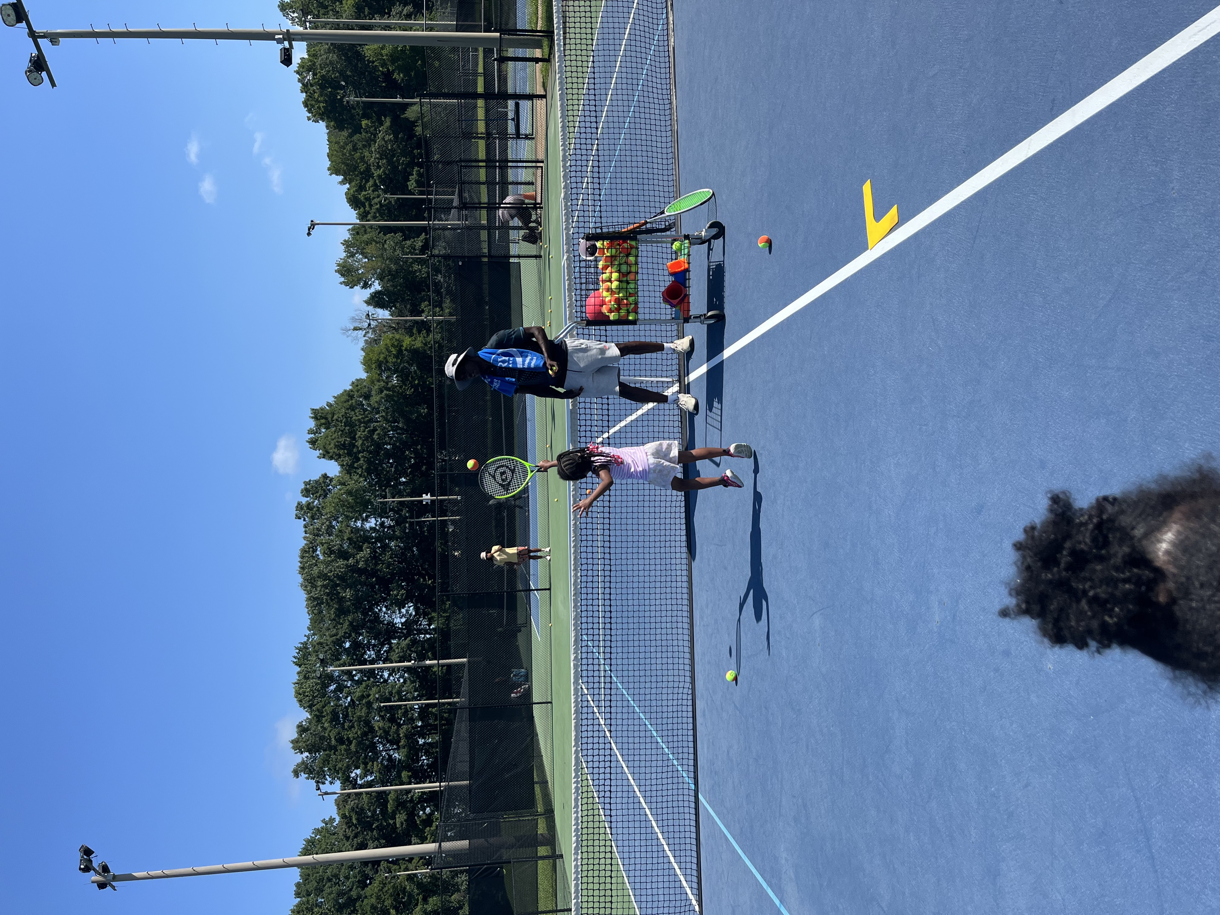 Coach Tee Reed leading a junior tennis lesson on an outdoor hard court in Roswell, GA, feeding balls from a hopper while a young player practices her groundstrokes.