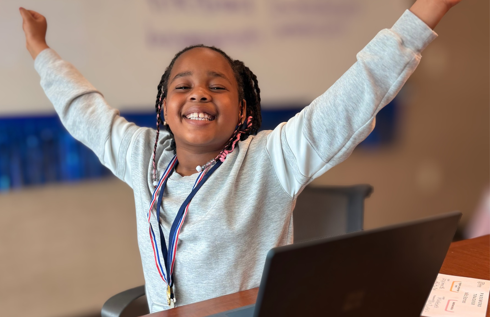 A smiling student wearing a medal raises her arms beside a laptop