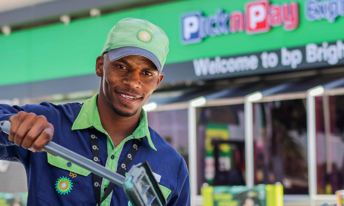 Male employee in navy and green uniform holding a squeegee in front of a Pick n Pay Express store.