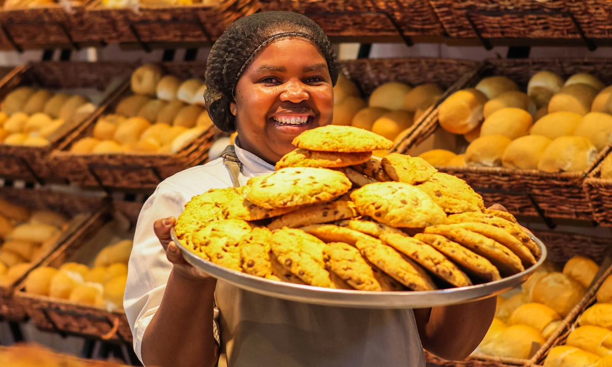 A smiling bakery worker wearing a hairnet and uniform holding a large tray of fresh chocolate chip cookies in front of display shelves filled with bread.