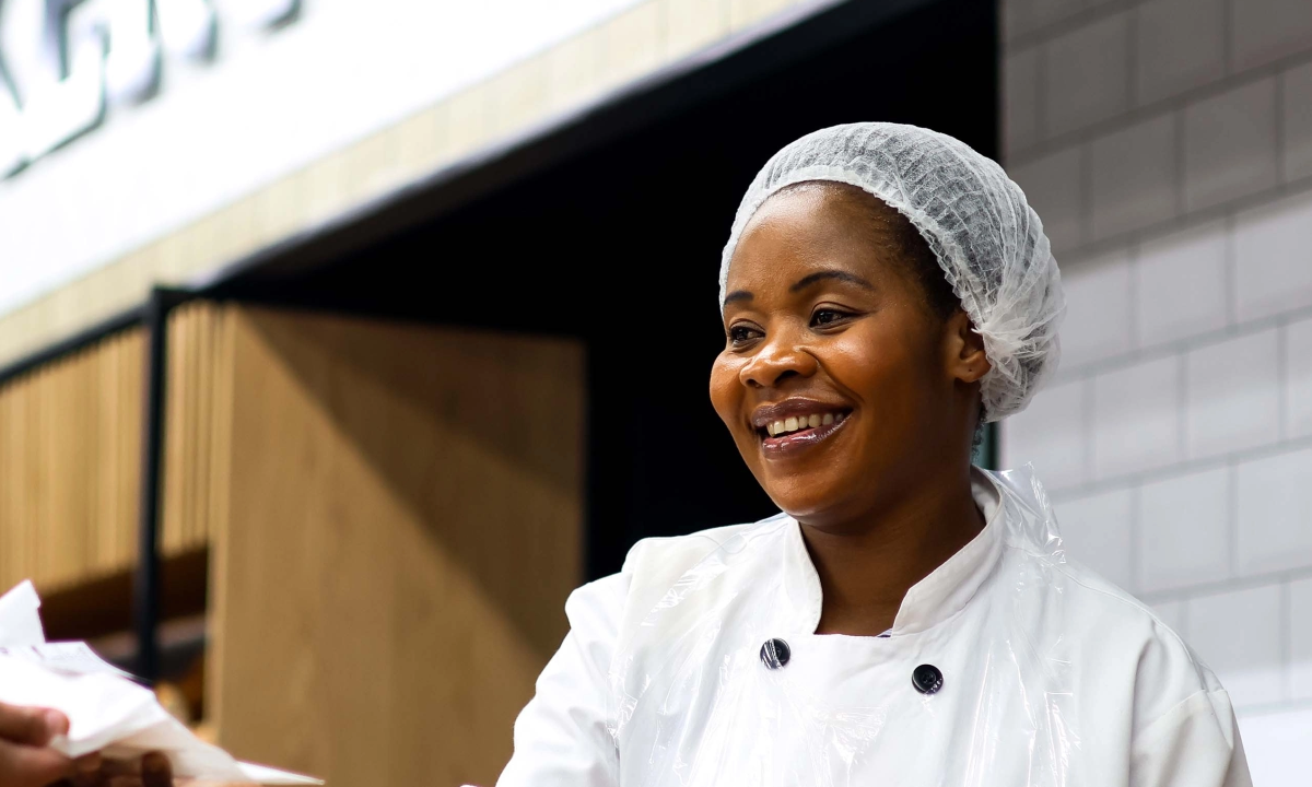 A smiling bakery worker in a white uniform and hairnet handing a package to a customer in a retail store.