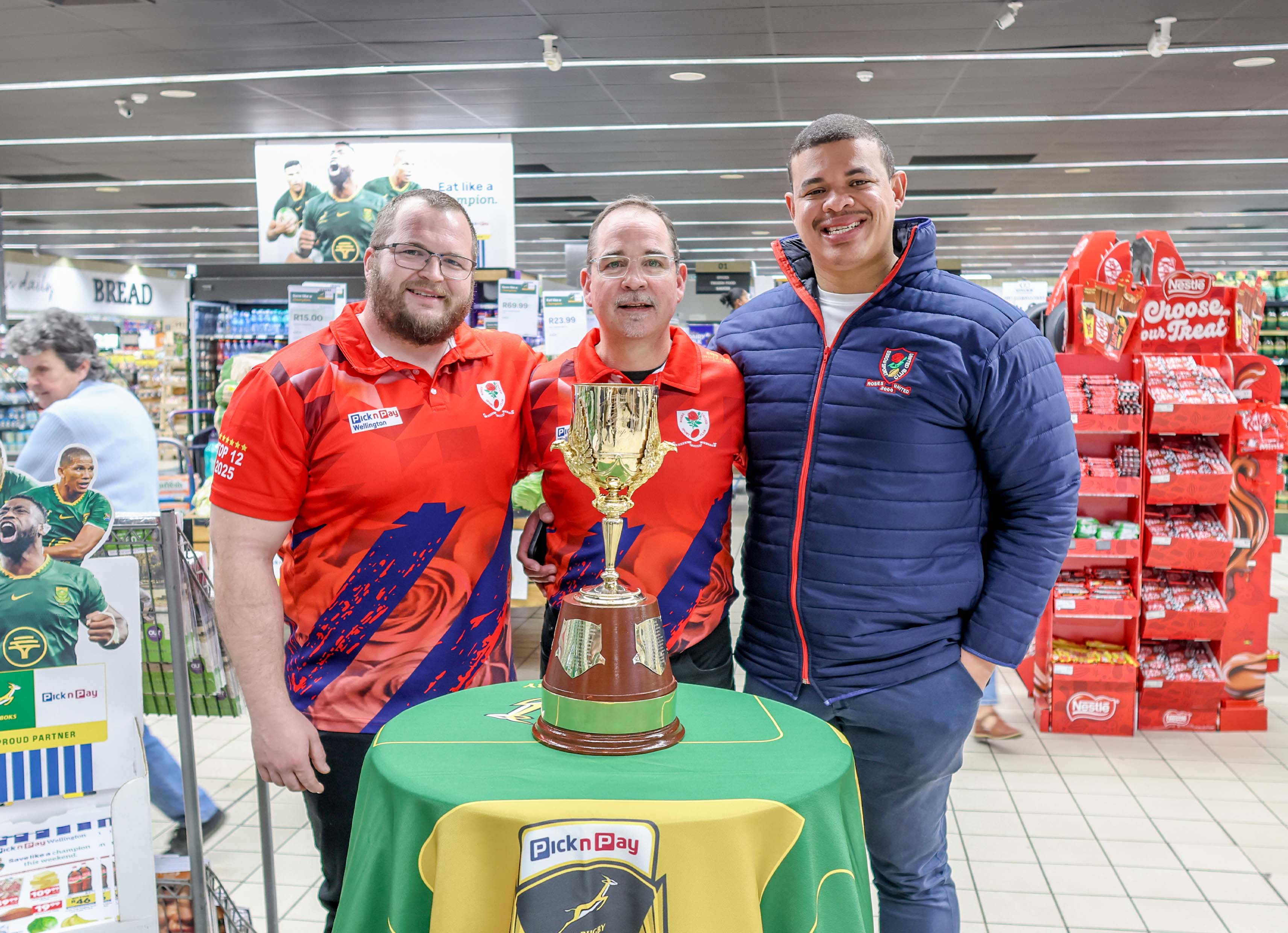 Three men stand behind a Pick n Pay table displaying the Gold Cup trophy inside the Pick n Pay Wellington store, with a Springbok promotional standee and an 'Eat like a champion' sign in the background.
