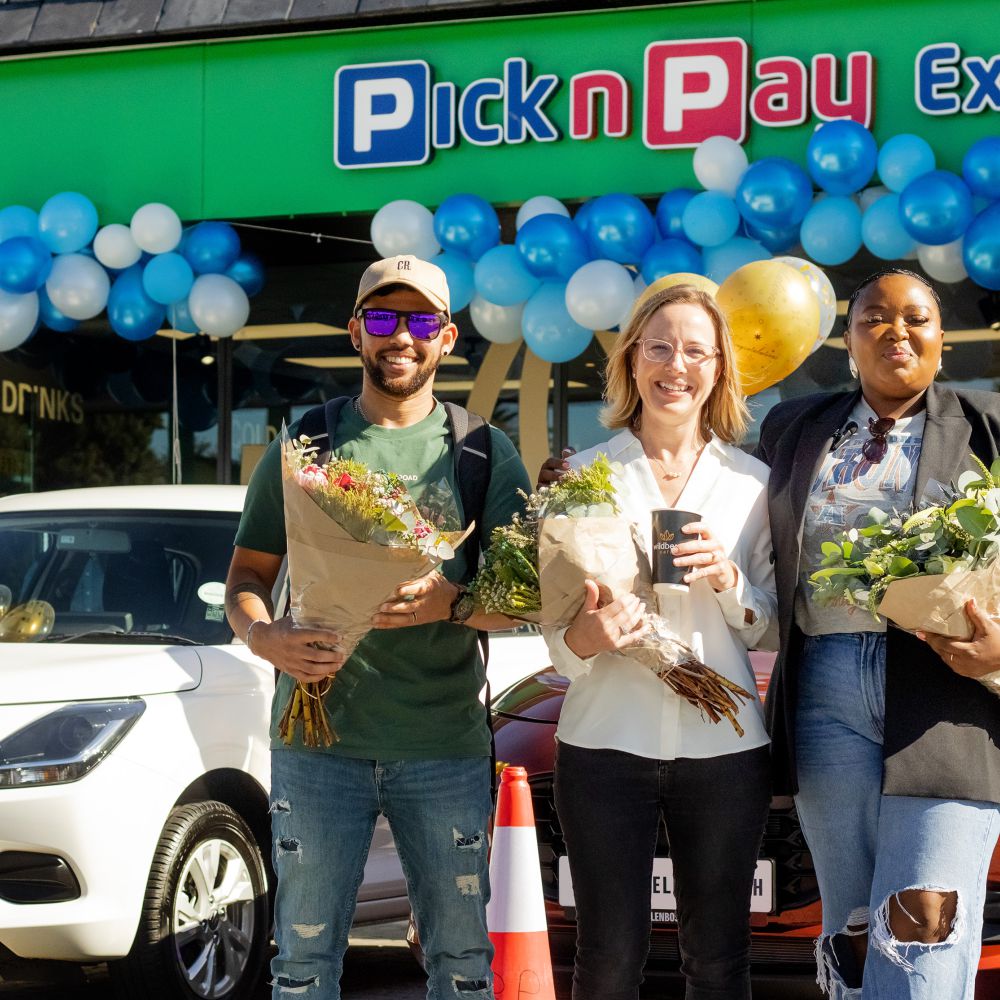 Three people holding flower bouquets pose in front of a Pick n Pay Express storefront decorated with blue and white balloons for a prize handover celebration.