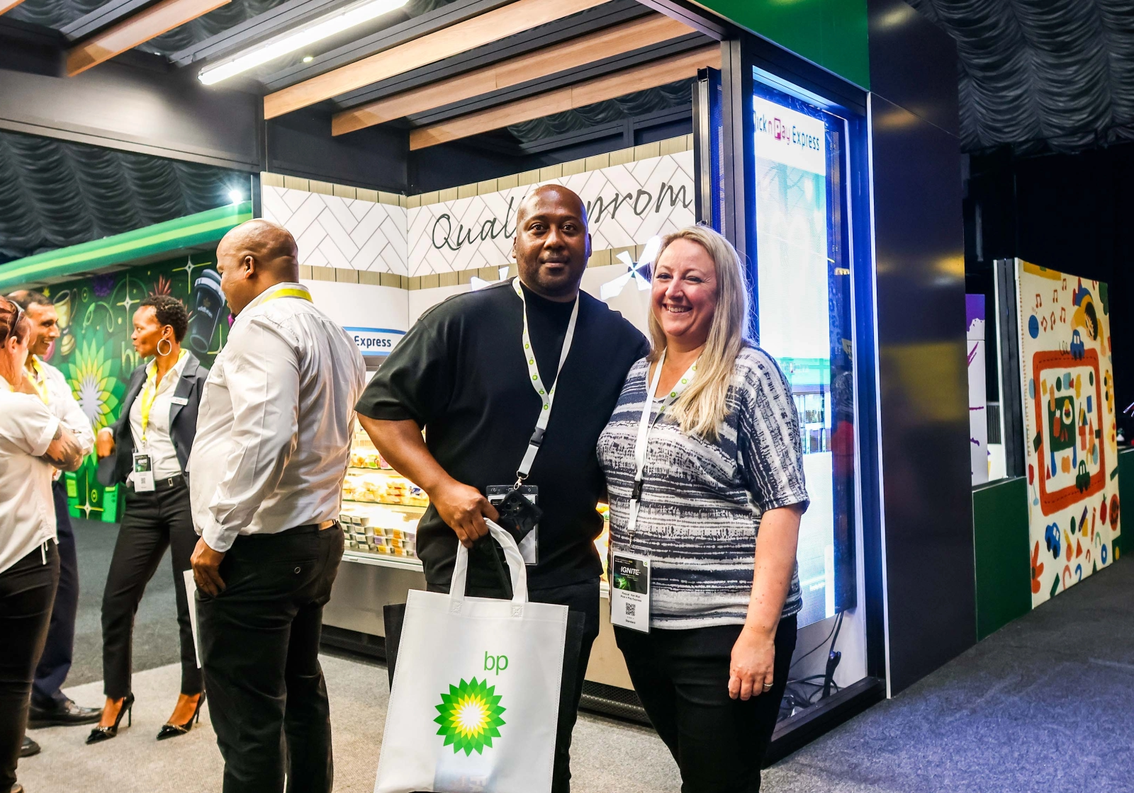 Two individuals posing in front of a branded Pick n Pay Express trade show booth at a corporate event, one holding a bp-branded tote bag.