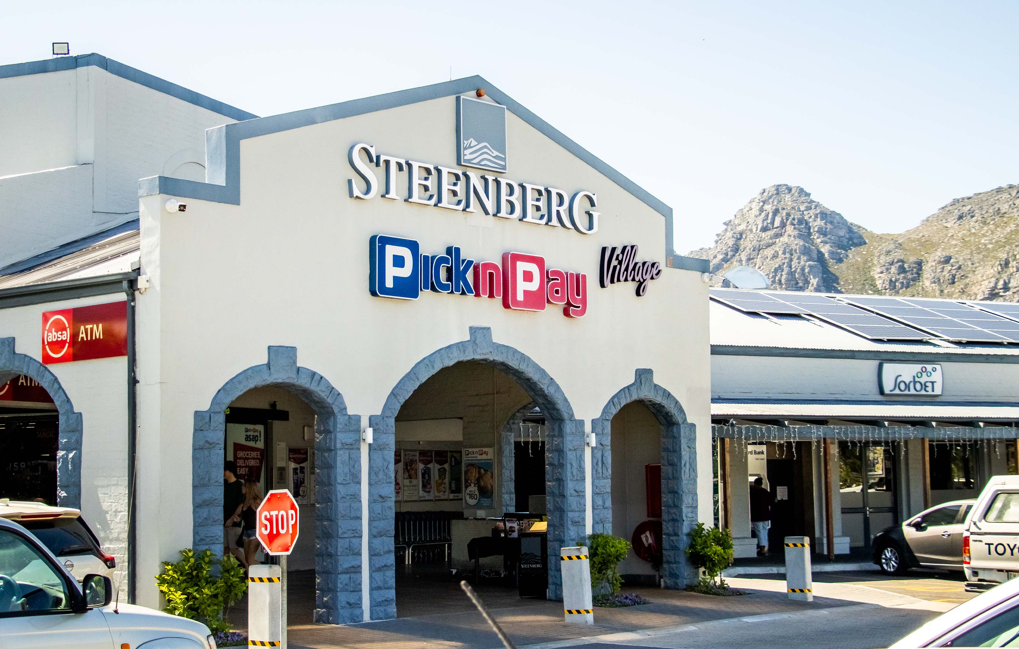 Exterior view of the Pick n Pay store at Steenberg Village shopping center, featuring blue-gray stone archways, Pick n Pay Village signage, Absa and Sorbet storefronts, with a mountain backdrop under a clear sky.