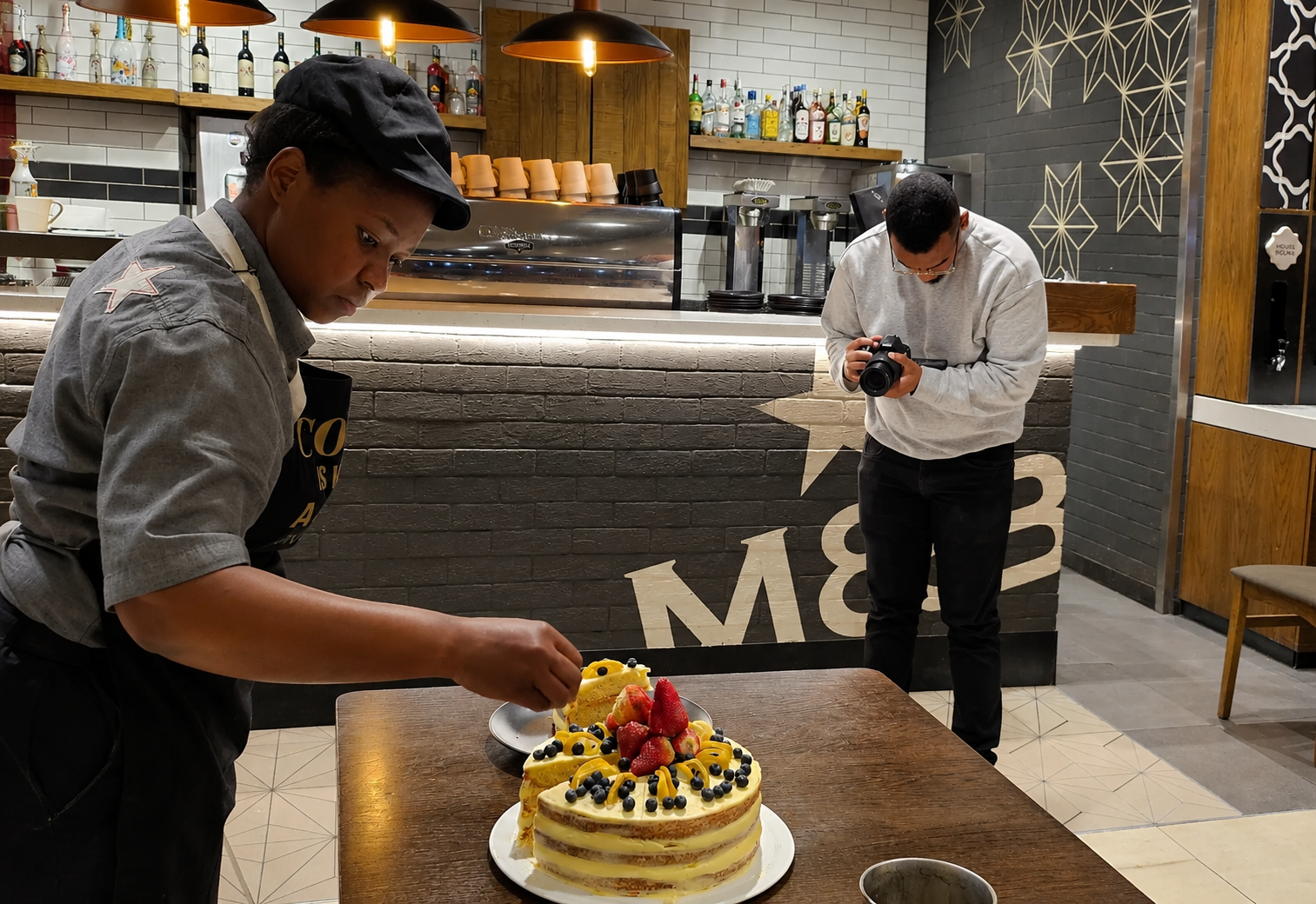 A food photoshoot in progress at a café: a staff member in a black apron styles a strawberry and blueberry layer cake on a wooden table while a photographer captures the shot with a DSLR camera.