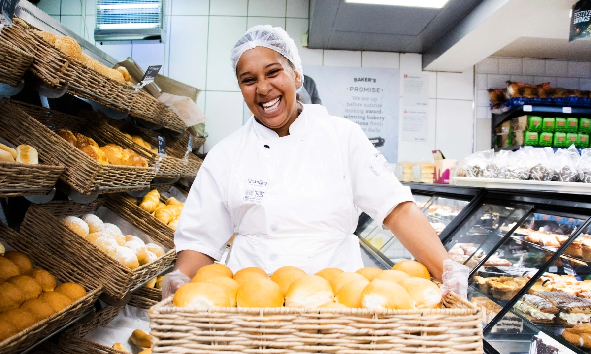 A smiling female bakery worker wearing a white uniform and hairnet, holding a large wicker basket of fresh bread rolls in a supermarket bakery aisle.