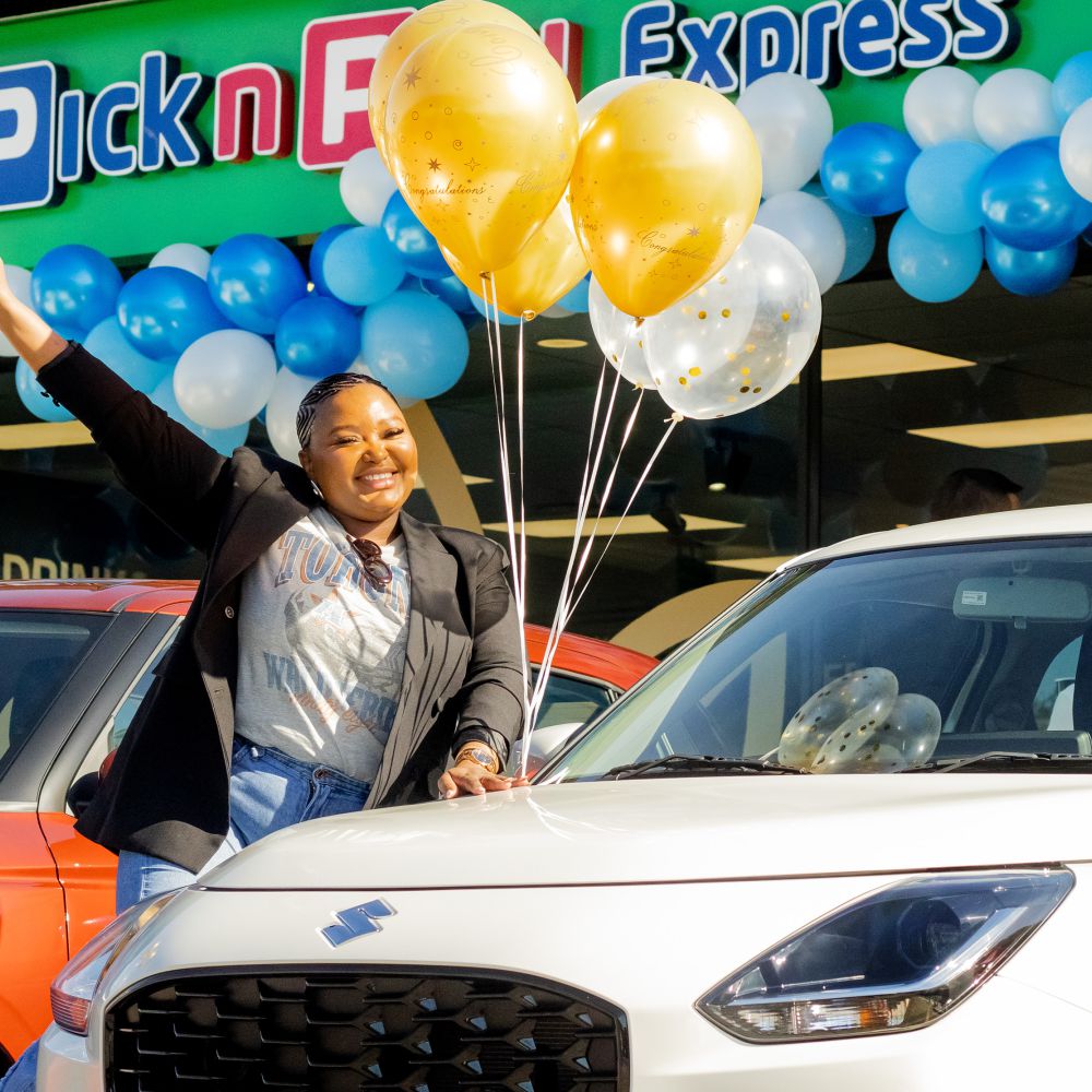 A smiling woman holding gold and confetti balloons poses next to a new white Suzuki car in front of a Pick n Pay Express store decorated with blue and white balloons for a prize handover.
