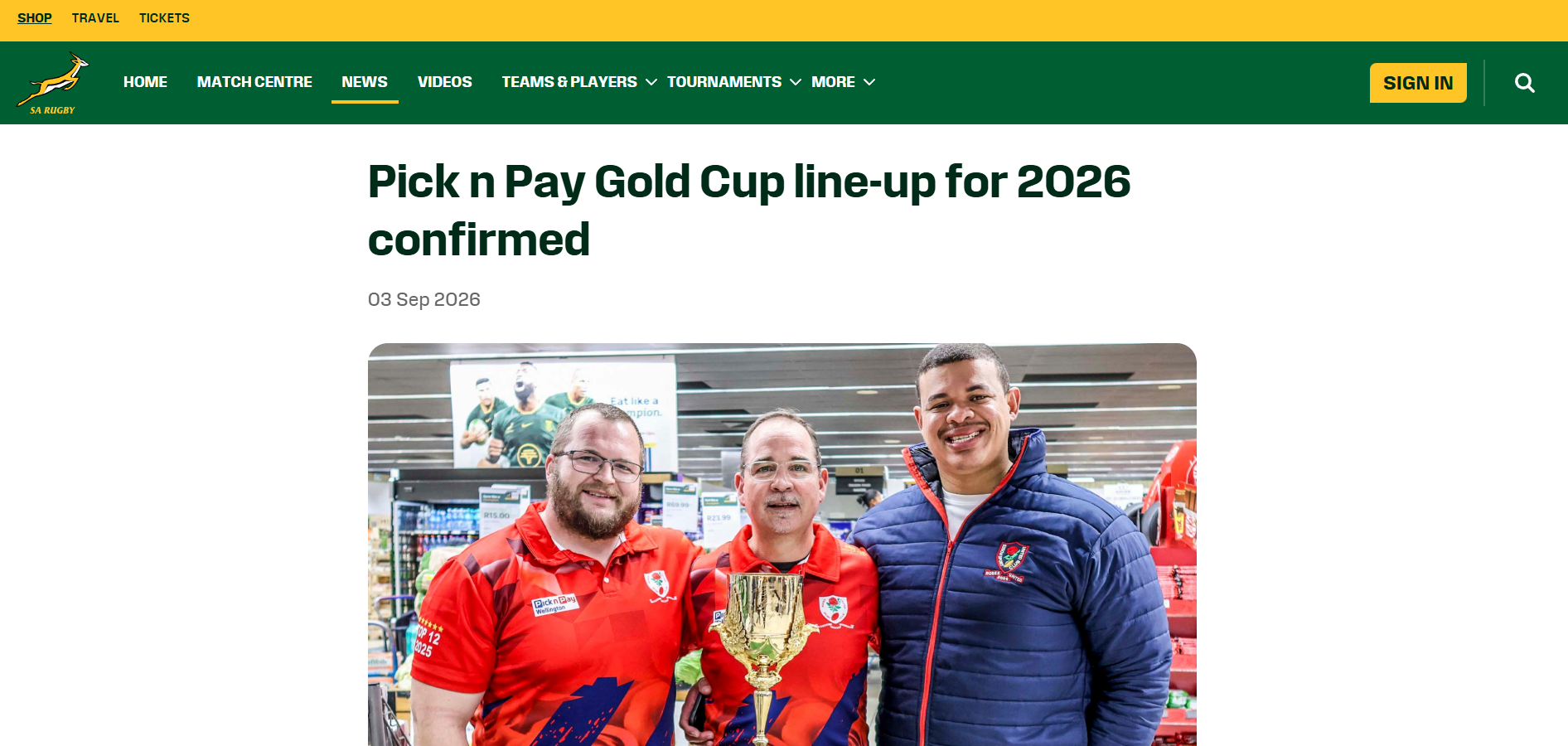 Three men stand together inside the Pick n Pay Wellington store, the centre figure holding the Gold Cup trophy.