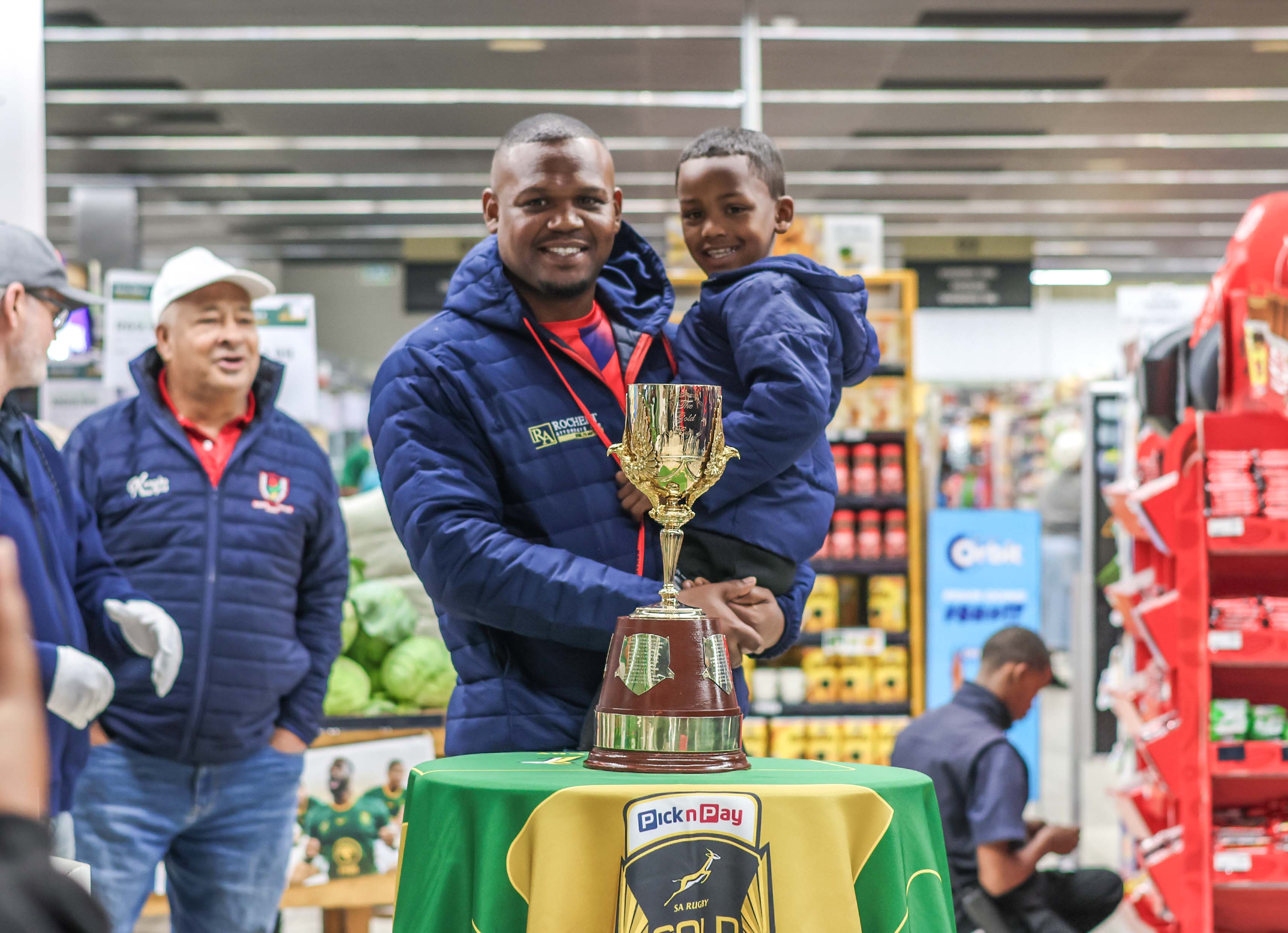 A man in a blue quilted jacket holds a smiling young boy at a Pick n Pay store event, with the Gold Cup trophy resting on a green and gold Pick n Pay display table in the foreground.