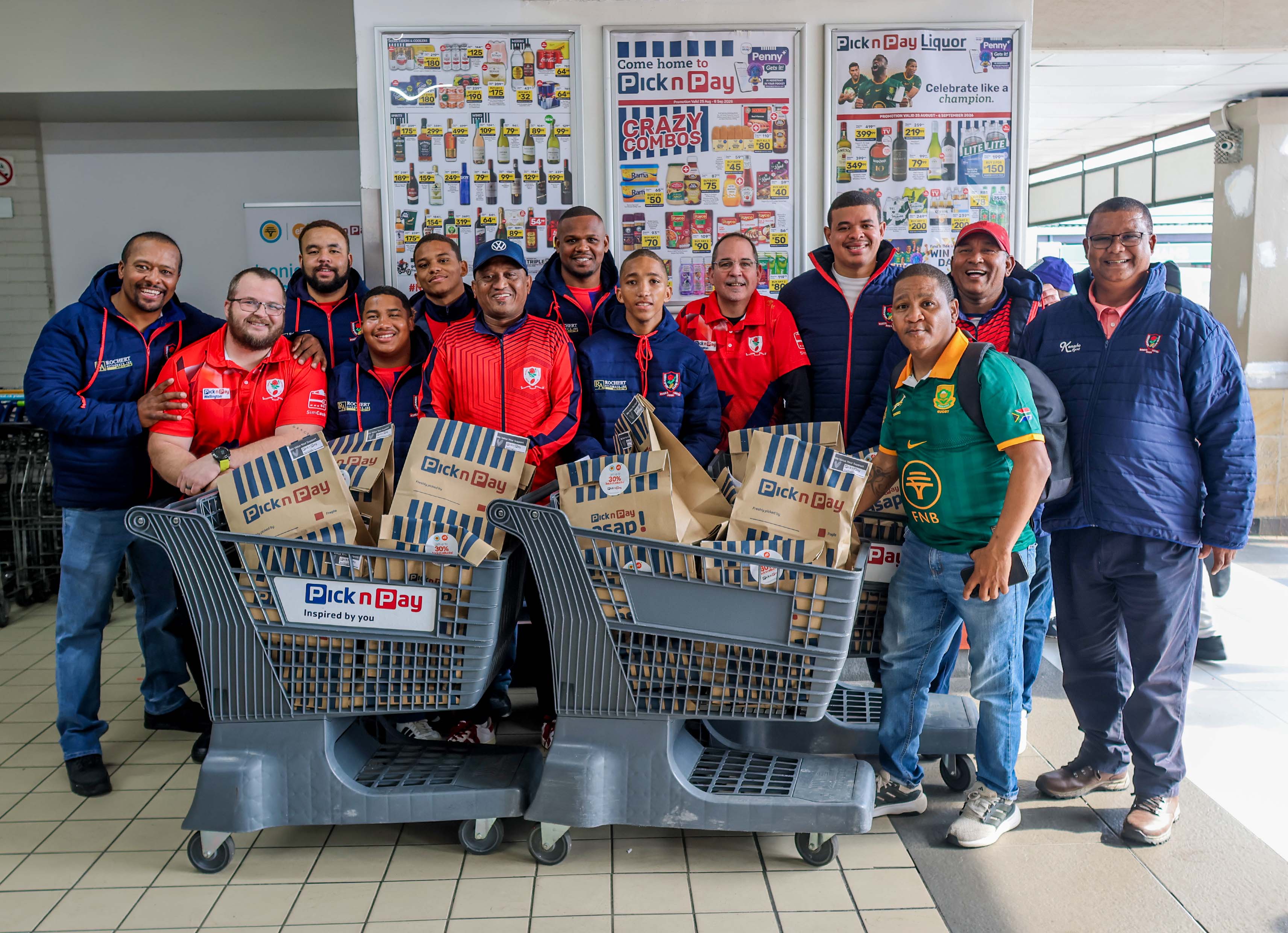 A group in Pick n Pay branded jackets and a green Springbok rugby jersey pose with two grocery carts filled with Pick n Pay paper bags in the Wellington store.