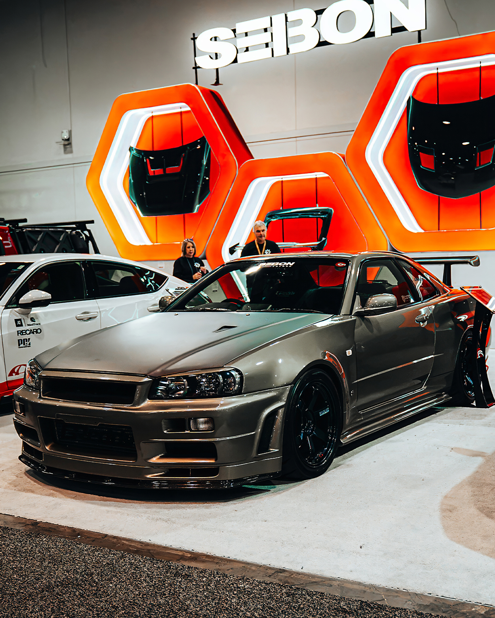 Seibon Carbon GT-R R34 - photography automotive photography by Chan2blazed Media: Nissan Skyline GT-R (R34) on display at the Seibon Carbon booth — metallic grey, carbon accents, and orange hexagonal backdrops.