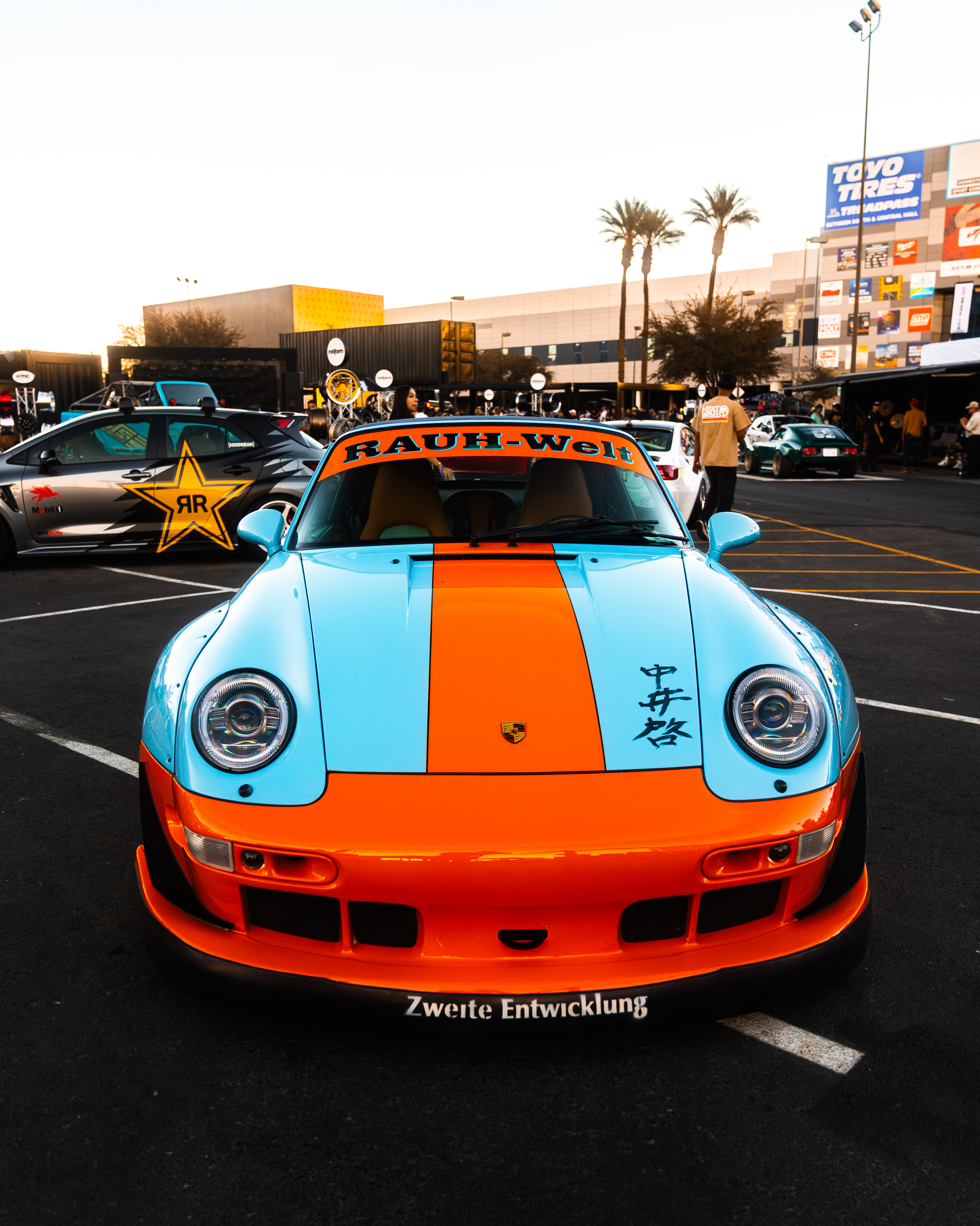 Porsche 911 RWB - photography automotive photography by Chan2blazed Media: A Rauh-Welt Begriff-built Porsche 911 at a golden-hour car event — sky-blue body with a vibrant orange racing stripe, RAUH-Welt windshield banner, and Japanese-character hood lettering.