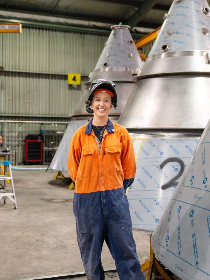 Young woman in a welding helmet and high-vis overalls smiling in a manufacturing workshop