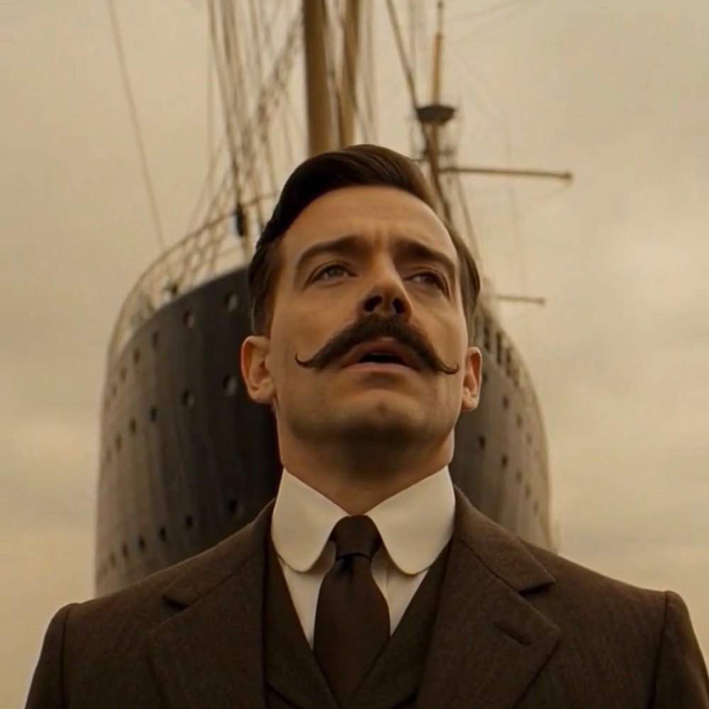 Film still of Charles Thomas, a man with a handlebar mustache in an Edwardian three-piece suit, in front of an ocean liner's bow.